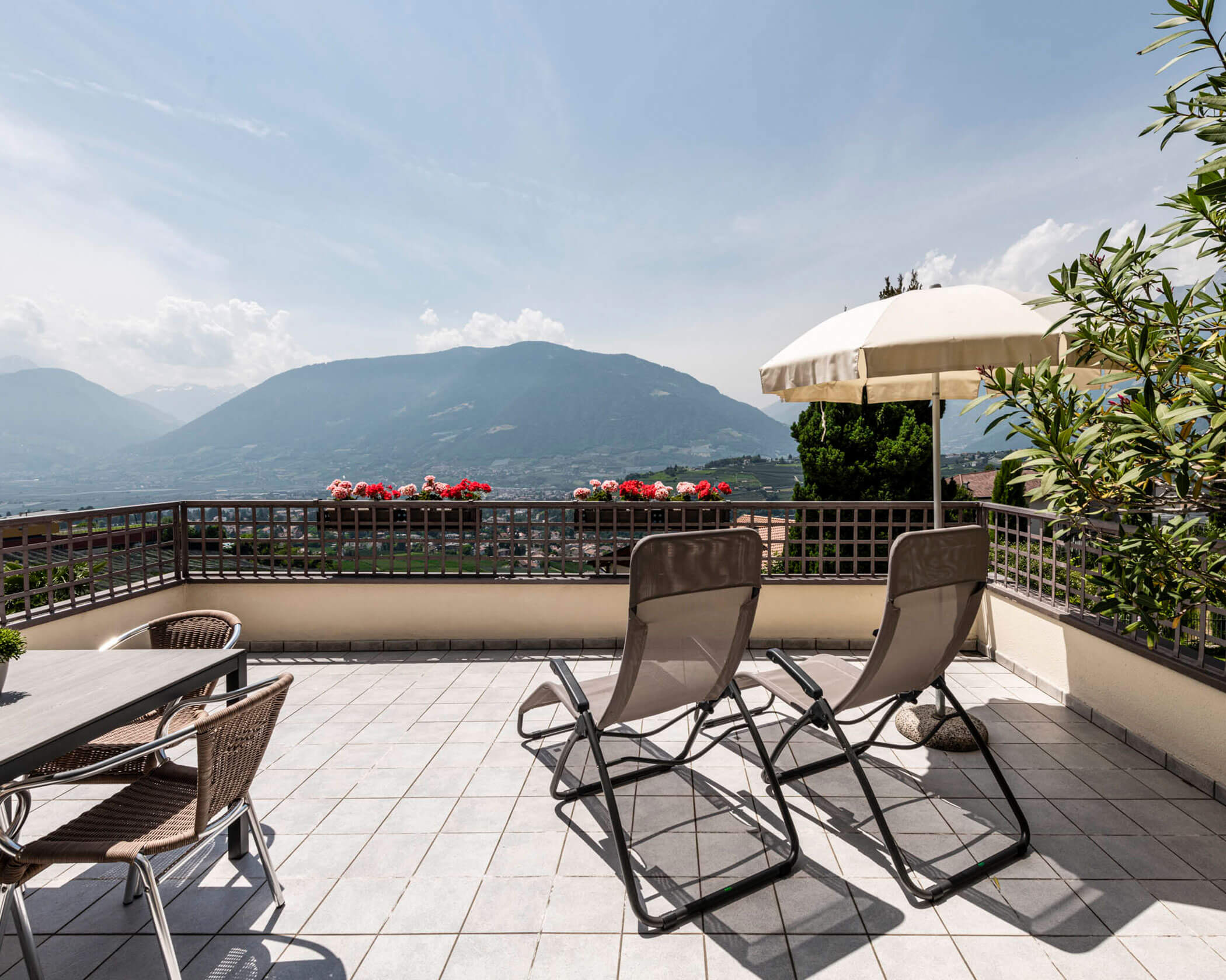 Two lounge chairs and a table on a sunny terrace with flowers, overlooking mountains and a valley. - Hotel Lagrein