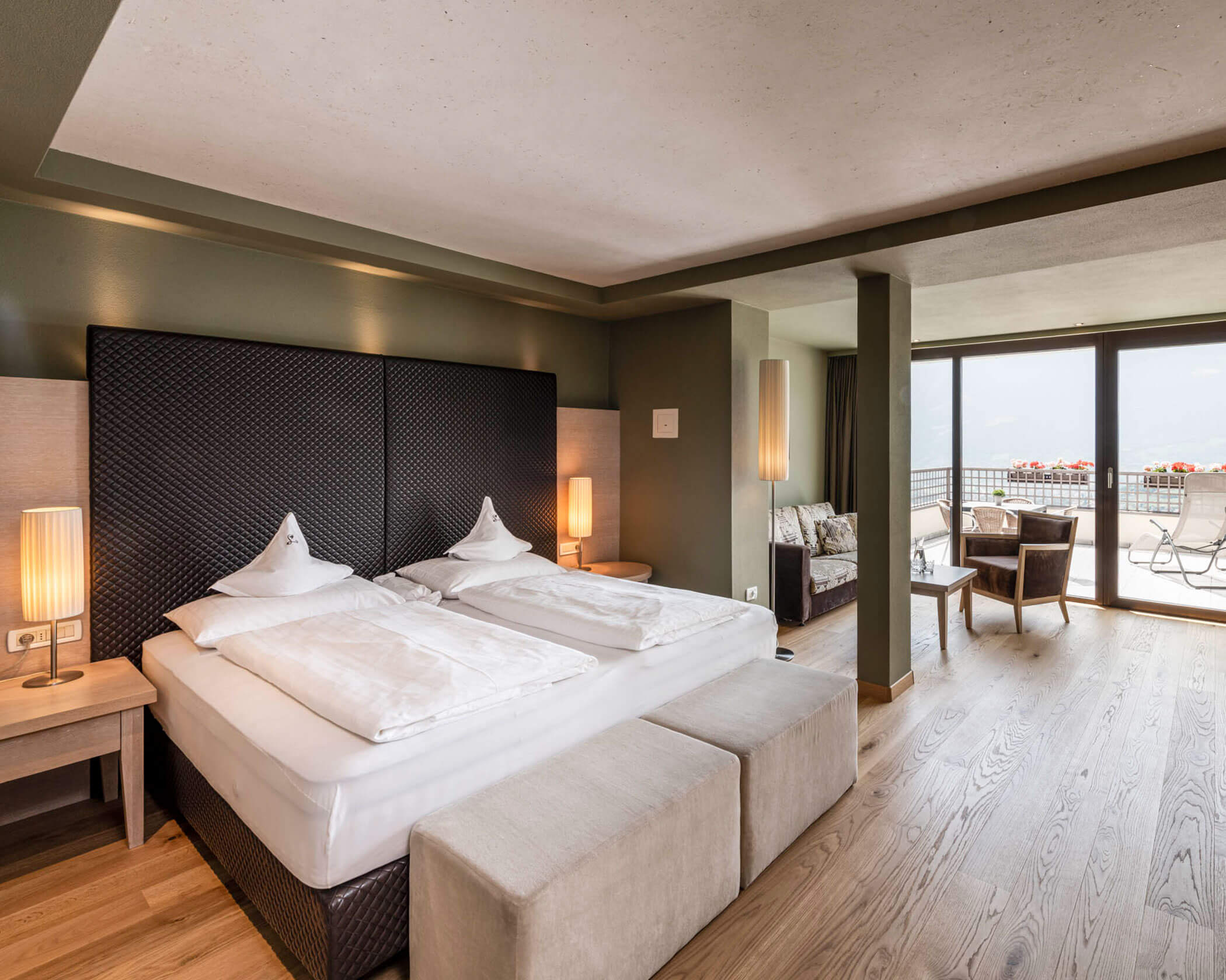 Modern hotel room with a large bed, seating area, and balcony with outdoor furniture and a scenic view. - Hotel Lagrein
