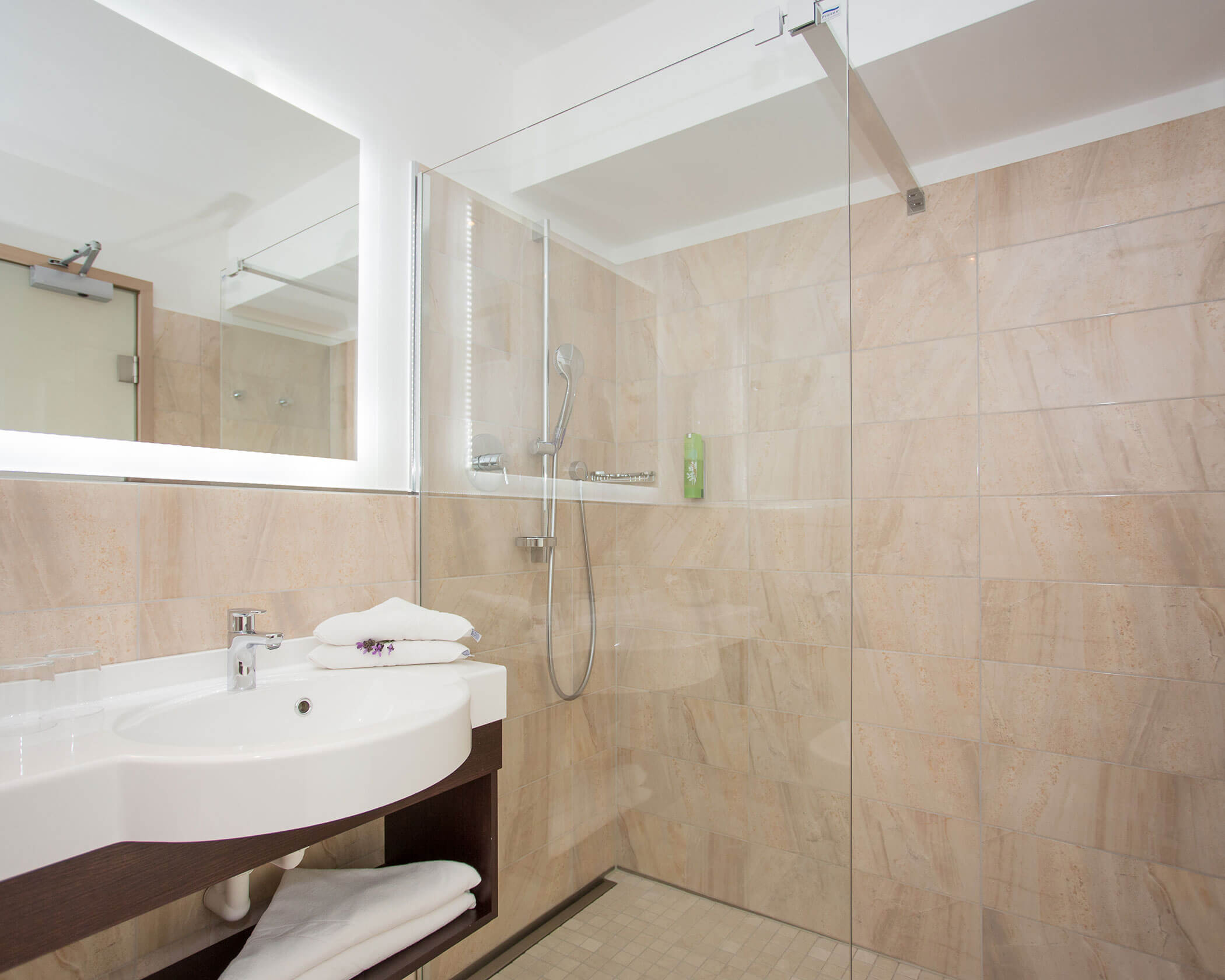 Modern bathroom with a glass shower, beige tiled walls, white sink, and neatly folded towels. - Hotel Lagrein