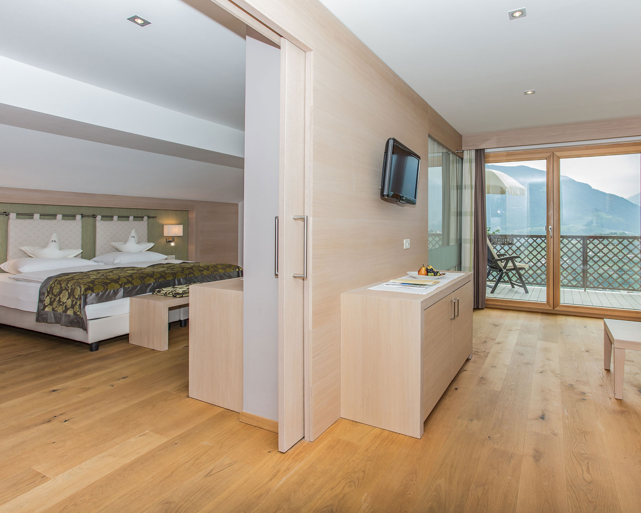 Modern hotel suite with wooden floors, a double bed, TV, balcony, and mountain view through glass doors. - Hotel Lagrein