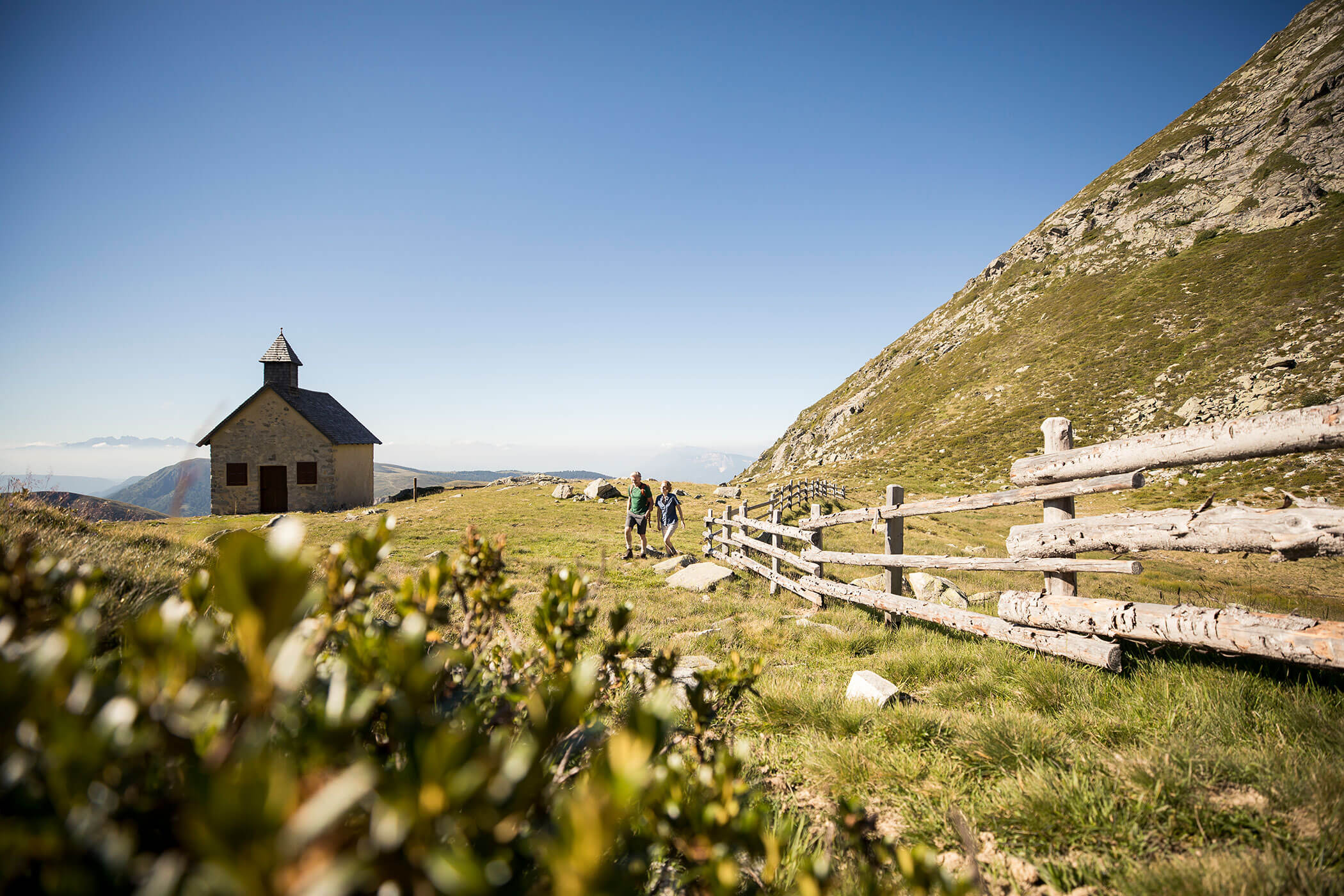 Small stone chapel and three people walking along a wooden fence in a grassy mountain landscape under a clear sky. - Hotel Lagrein