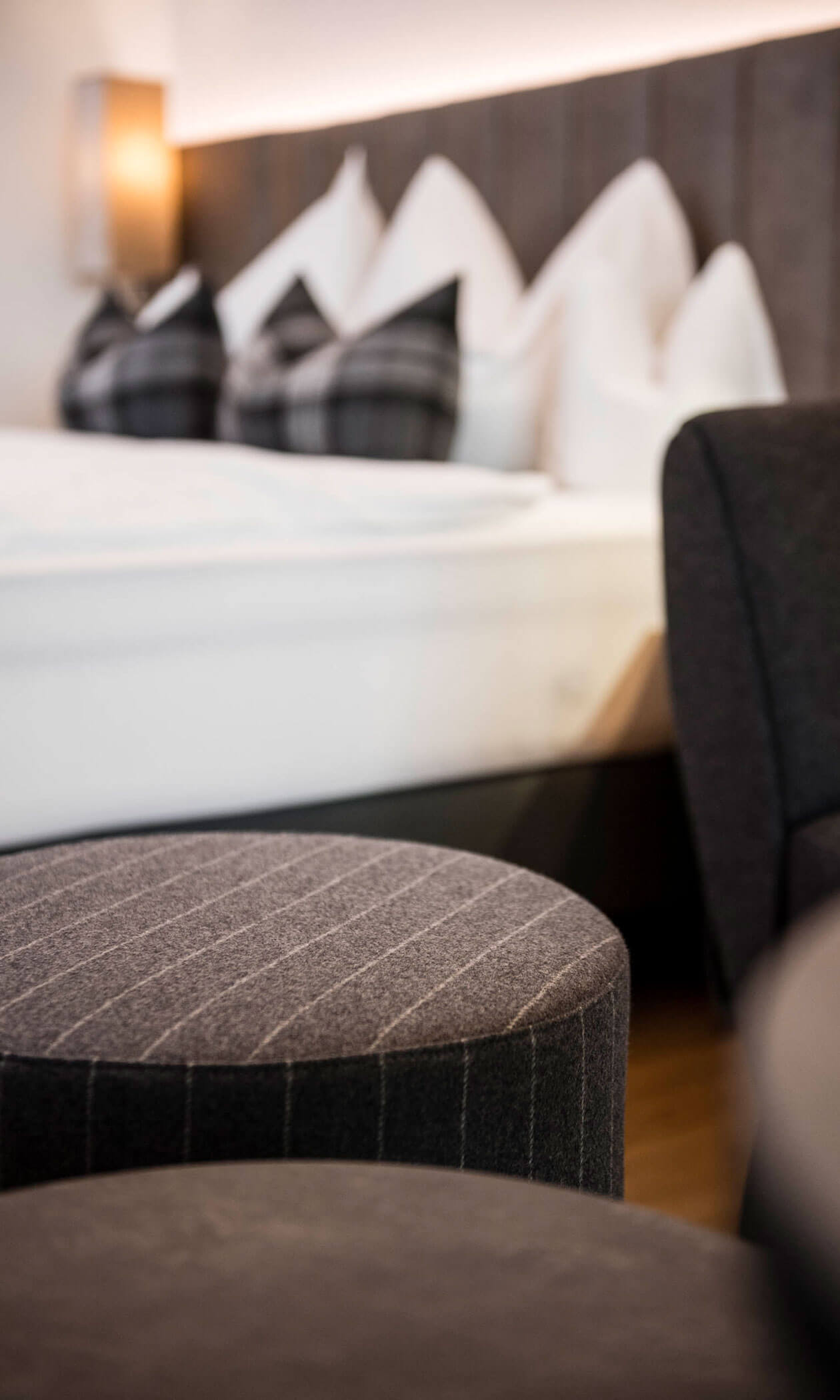 Close-up of a gray upholstered ottoman with a blurred bed and pillows in the background. - Hotel Lagrein
