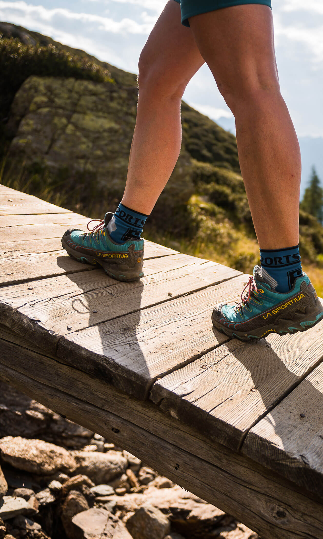 Person wearing teal hiking shoes and blue socks walking up a wooden bridge outdoors in sunlight. - Hotel Lagrein