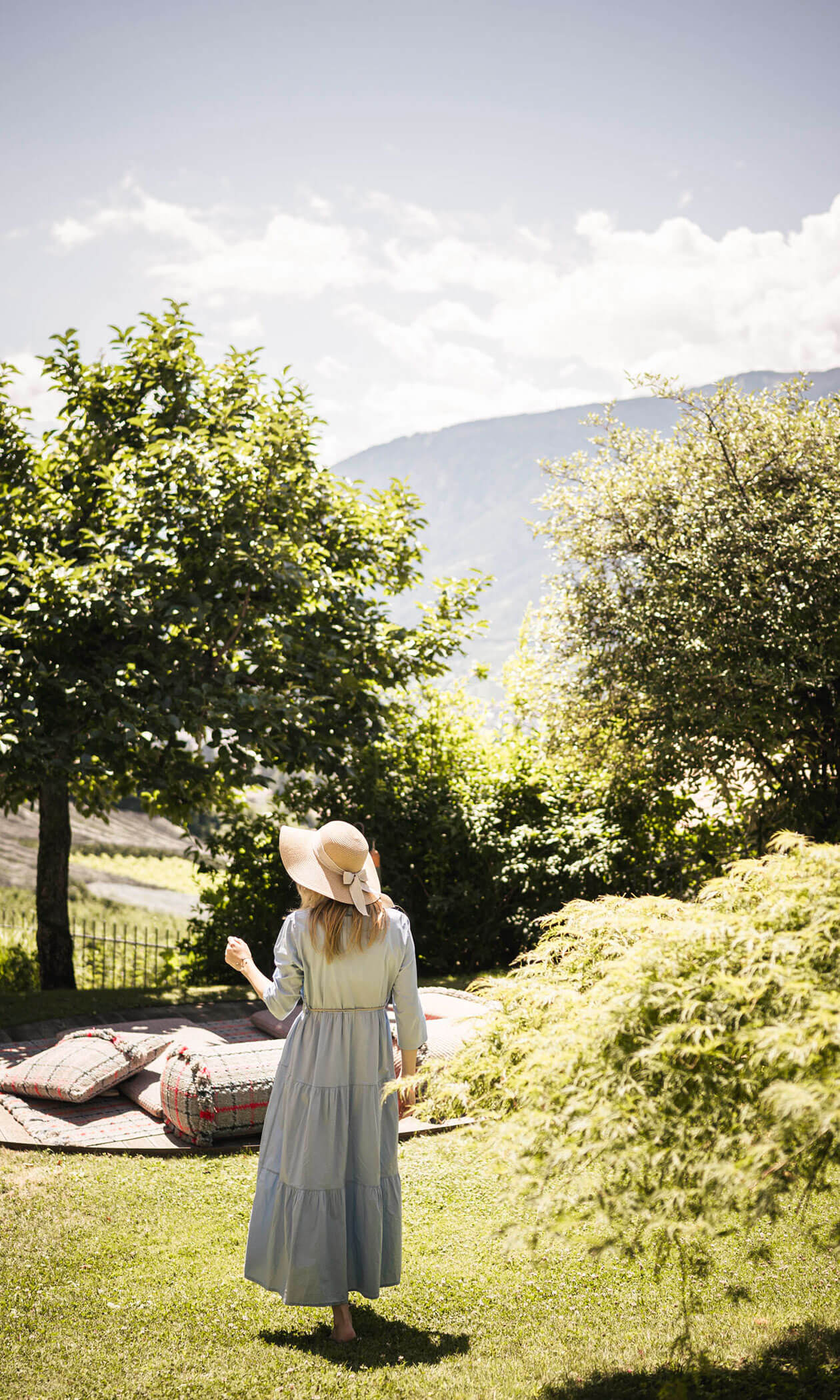 Woman in a light blue dress and sunhat walks barefoot on grass in a sunny, green garden with mountains in background. - Hotel Lagrein