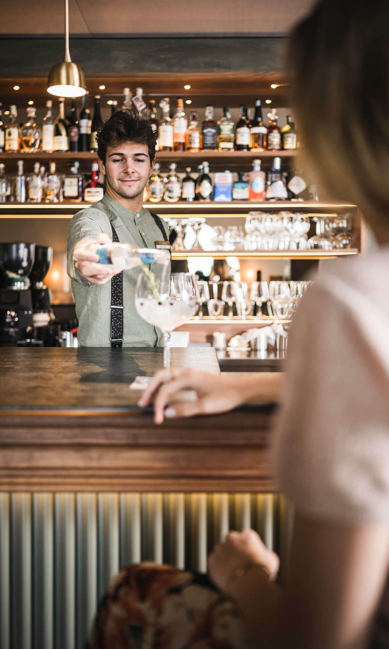 Bartender pouring a drink for a customer seated at a bar with shelves of bottles in the background. - Hotel Lagrein