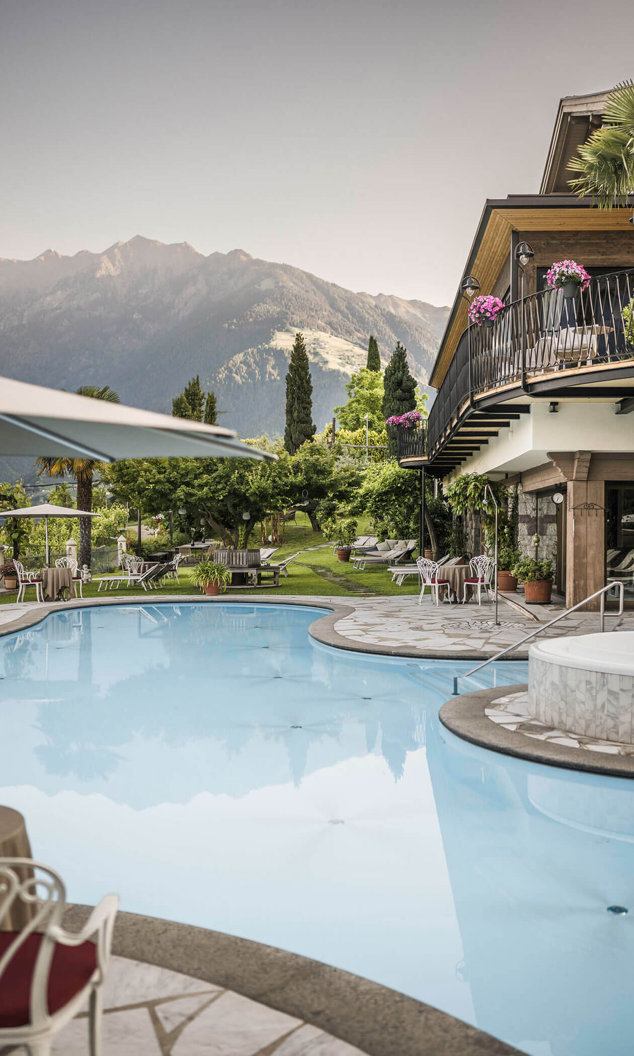 Outdoor pool with lounge chairs, greenery, and mountains in the background on a clear day. - Hotel Lagrein