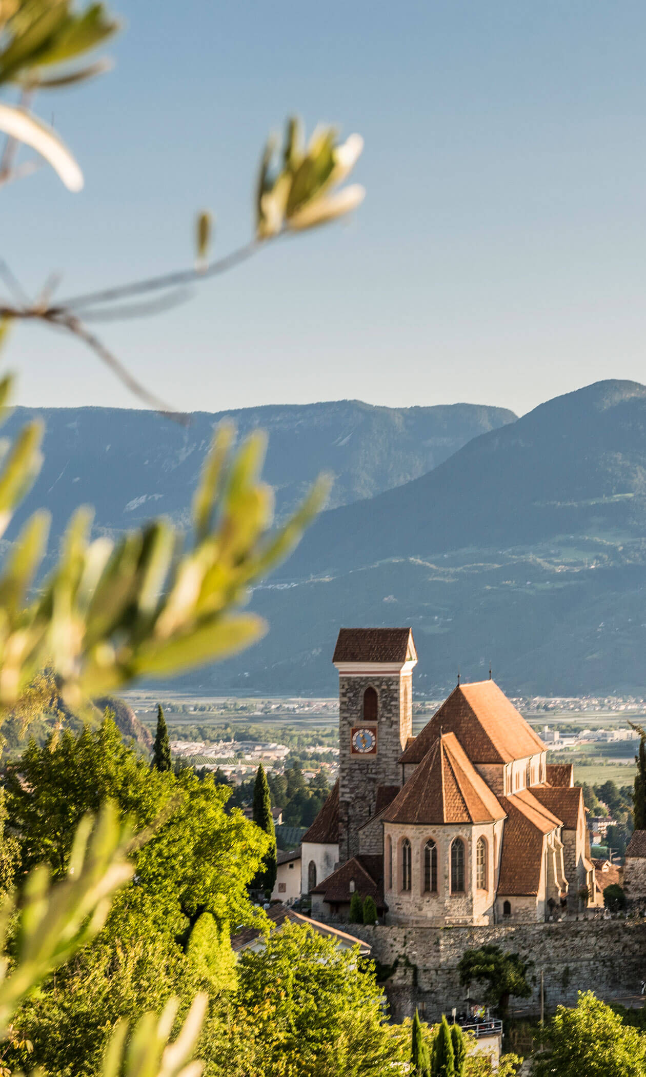 A stone church with a clock tower and red roof stands among greenery, with mountains in the background. - Hotel Lagrein