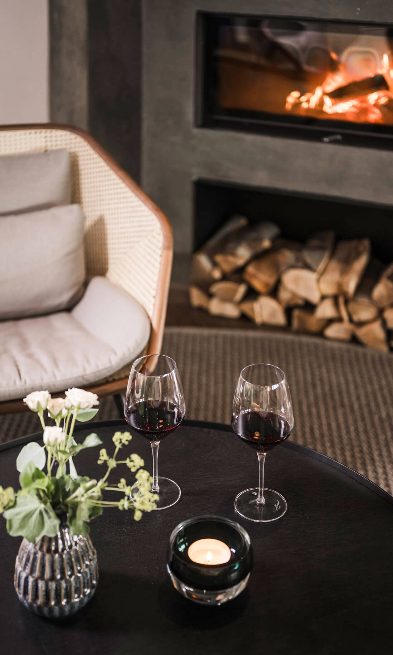 Two glasses of red wine on a table near a lit fireplace, with a chair and flowers in the background. - Hotel Lagrein