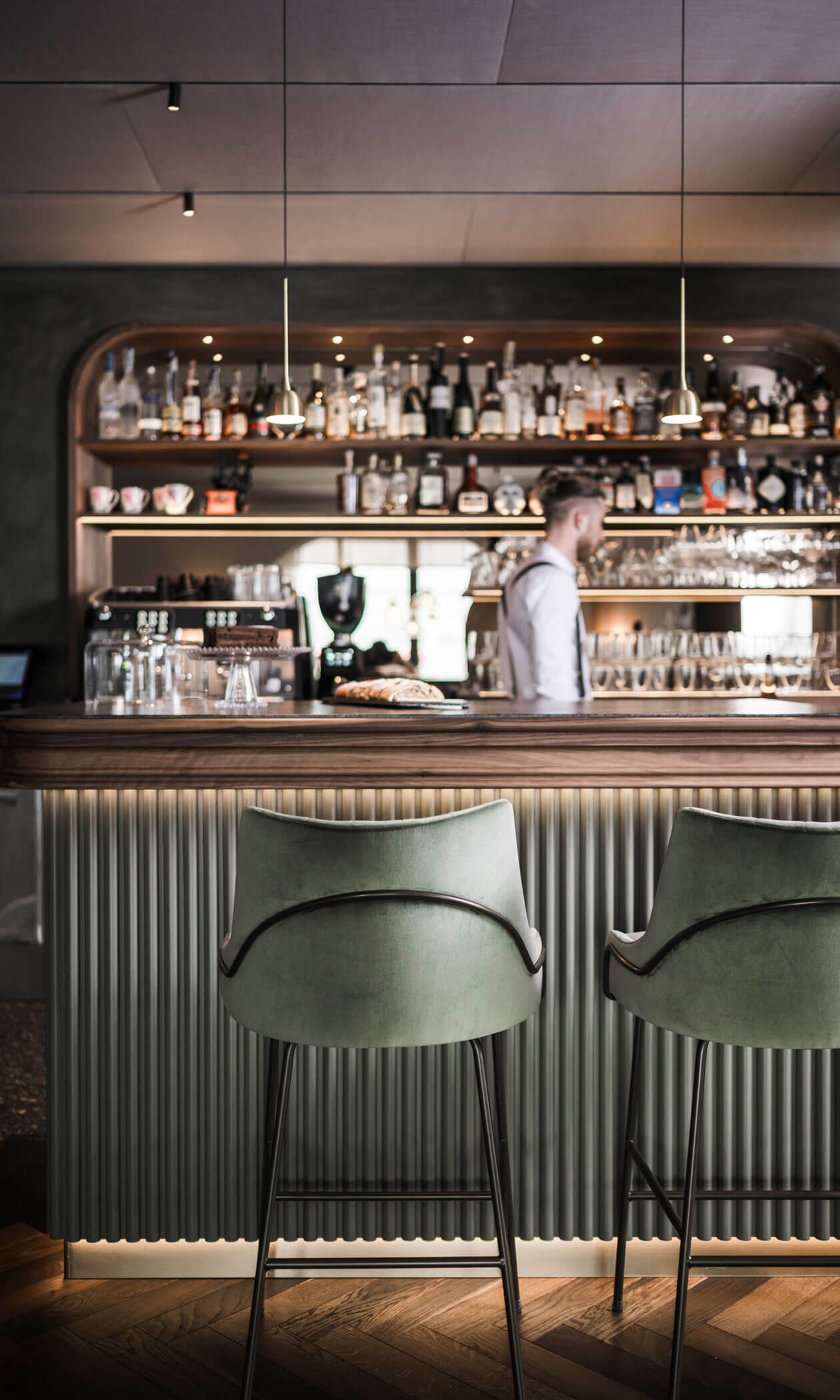 Two green barstools at a stylish bar with shelves of bottles and a bartender in the background. - Hotel Lagrein