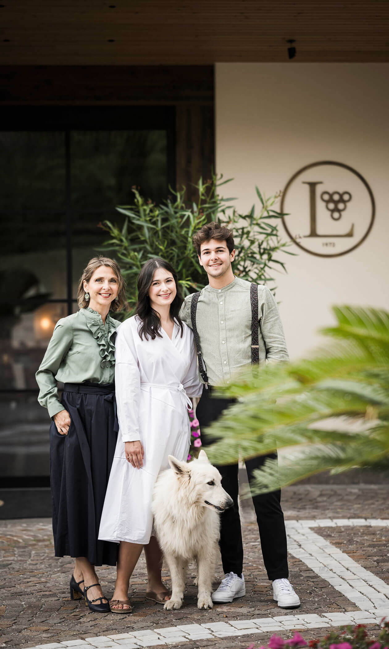 Three people stand smiling with a white dog outside a building, plants and a round logo are visible behind them. - Hotel Lagrein