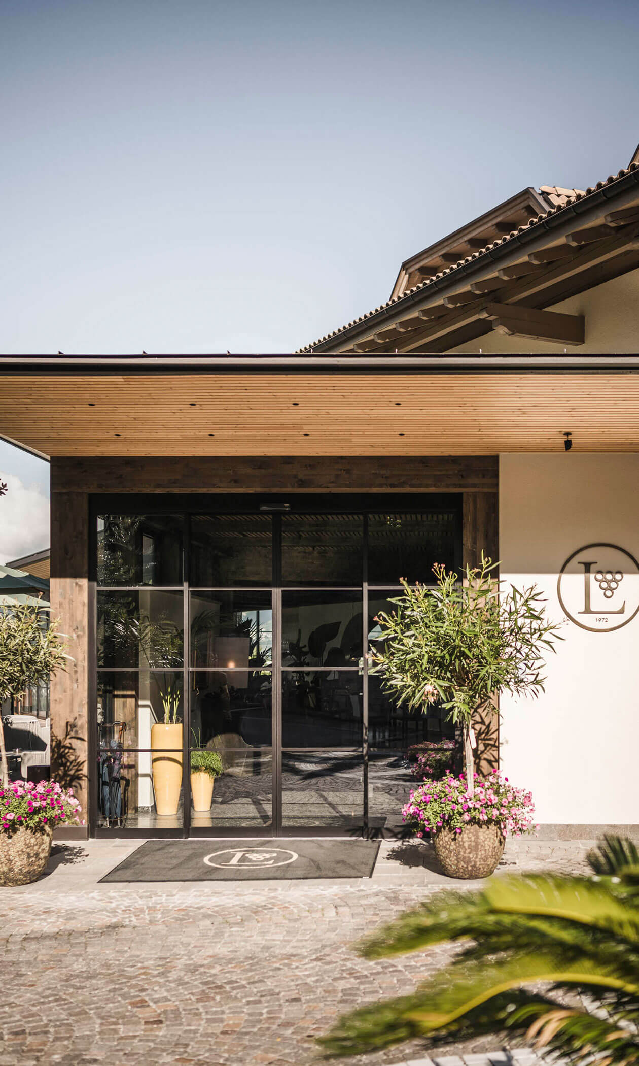 Modern building entrance with glass doors, potted plants, and a circular vineyard logo on the wall. - Hotel Lagrein