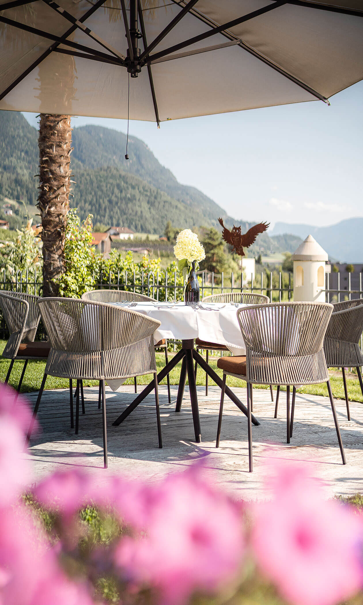 Outdoor dining table with flowers under a large umbrella, mountains and greenery in the background. - Hotel Lagrein