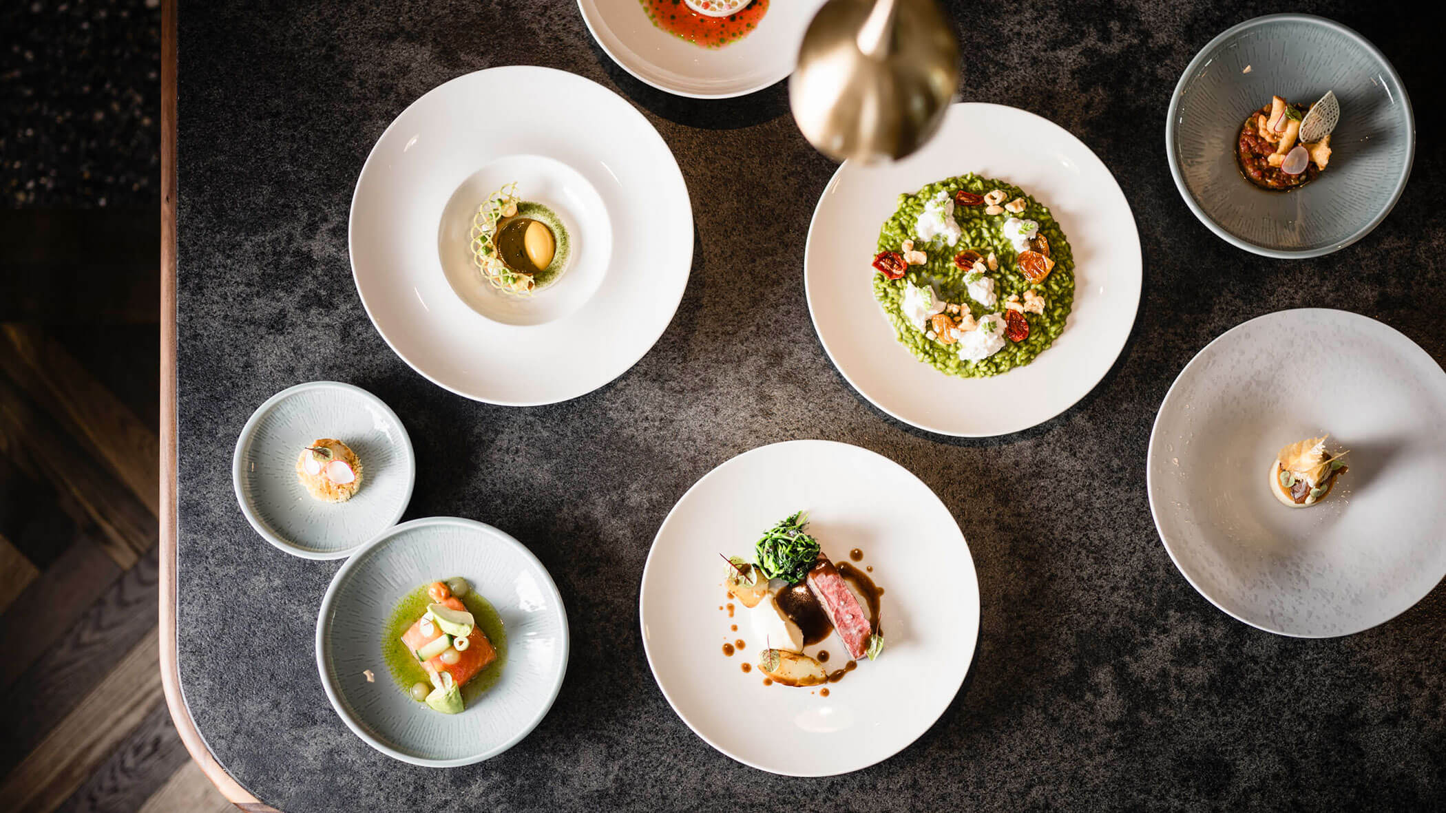 Overhead view of assorted gourmet dishes artfully plated on white and light blue plates on a dark table. - Hotel Lagrein
