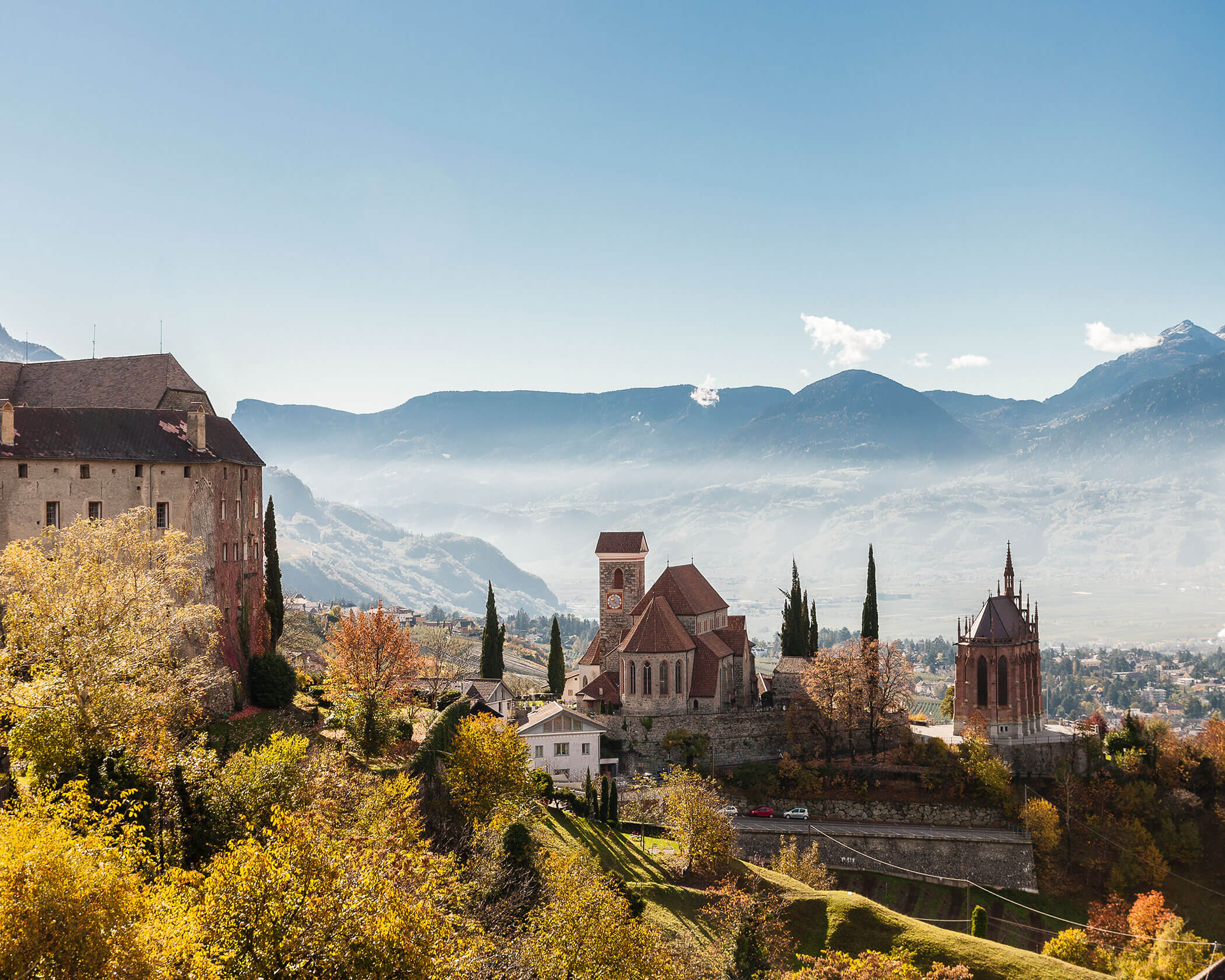 Hilltop village with stone buildings, towers, and autumn trees, set against distant blue mountains under a clear sky. - Hotel Lagrein