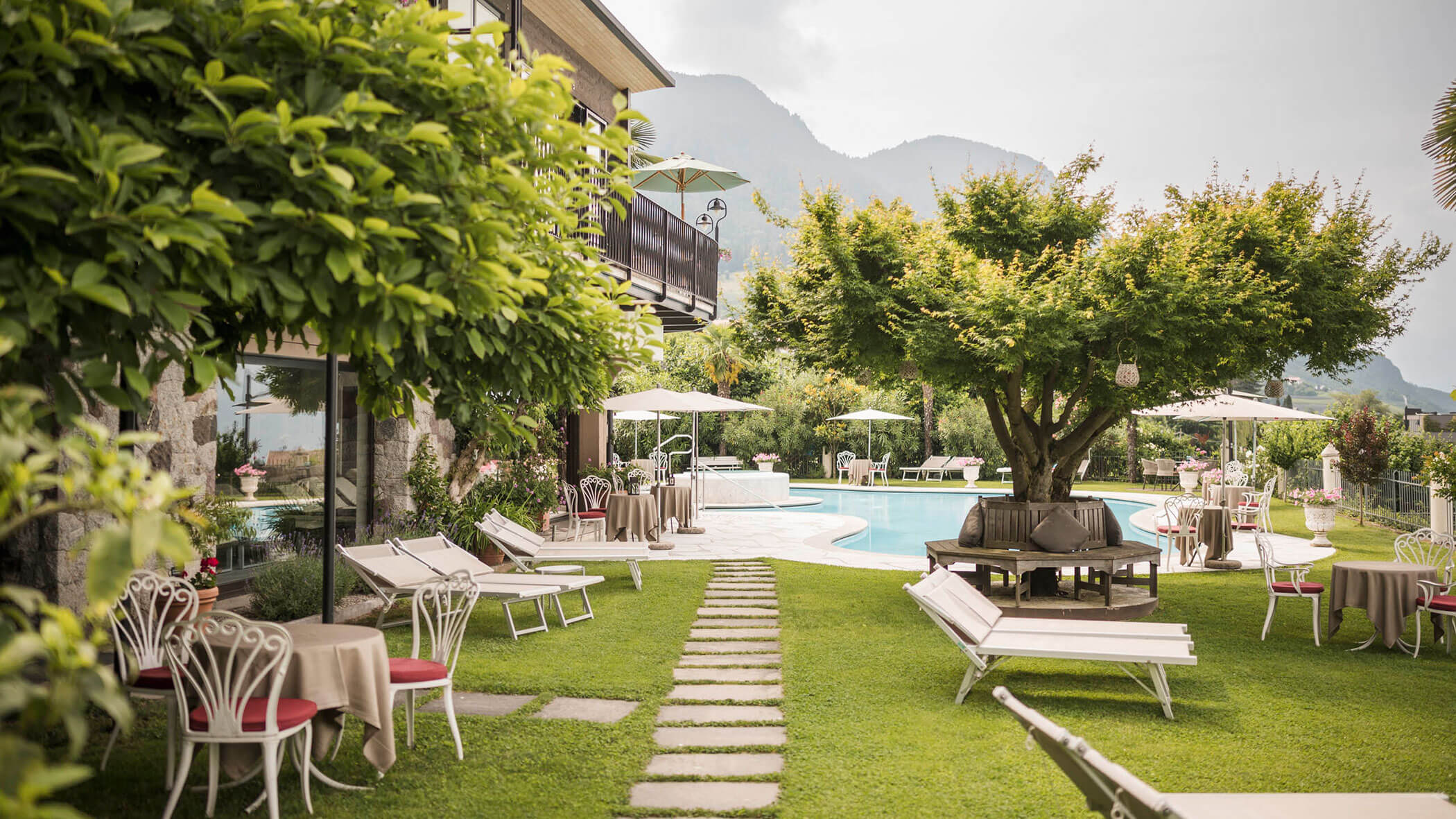 Sunny garden with tables, chairs, pool, and large trees, set against a backdrop of mountains. - Hotel Lagrein