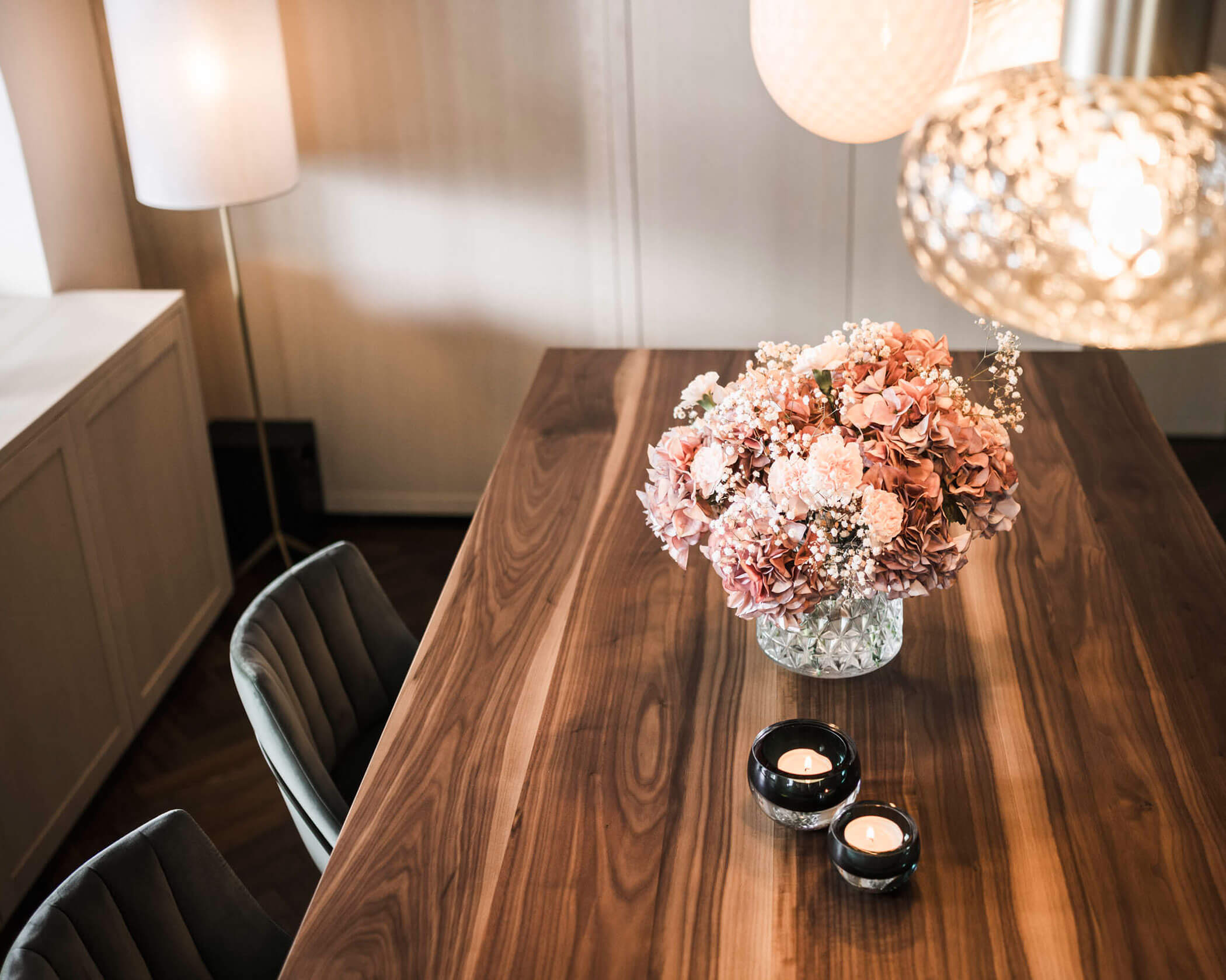 A wooden dining table with pink flowers in a vase and two lit candles in black holders. - Hotel Lagrein
