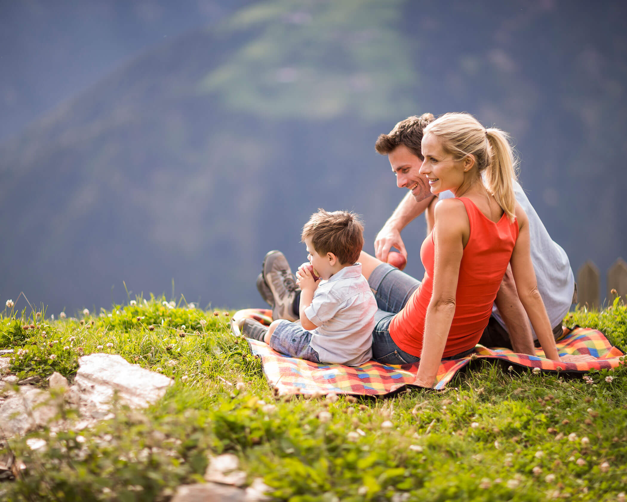 A family of three sits on a blanket outdoors, enjoying a scenic mountain view on a sunny day. - Hotel Lagrein