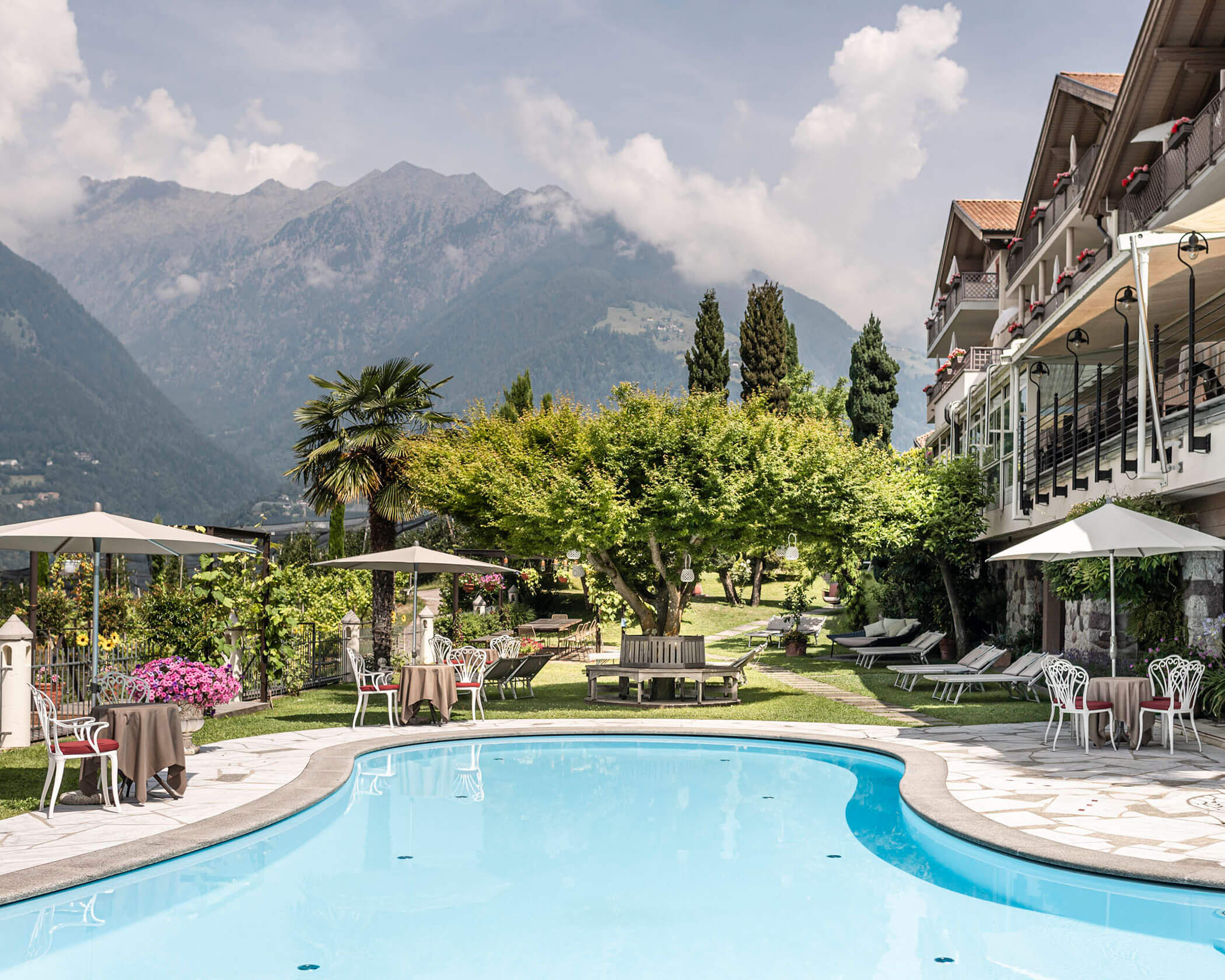 Outdoor pool with lounge chairs, umbrellas, and garden, set against mountain and hotel building backdrop. - Hotel Lagrein