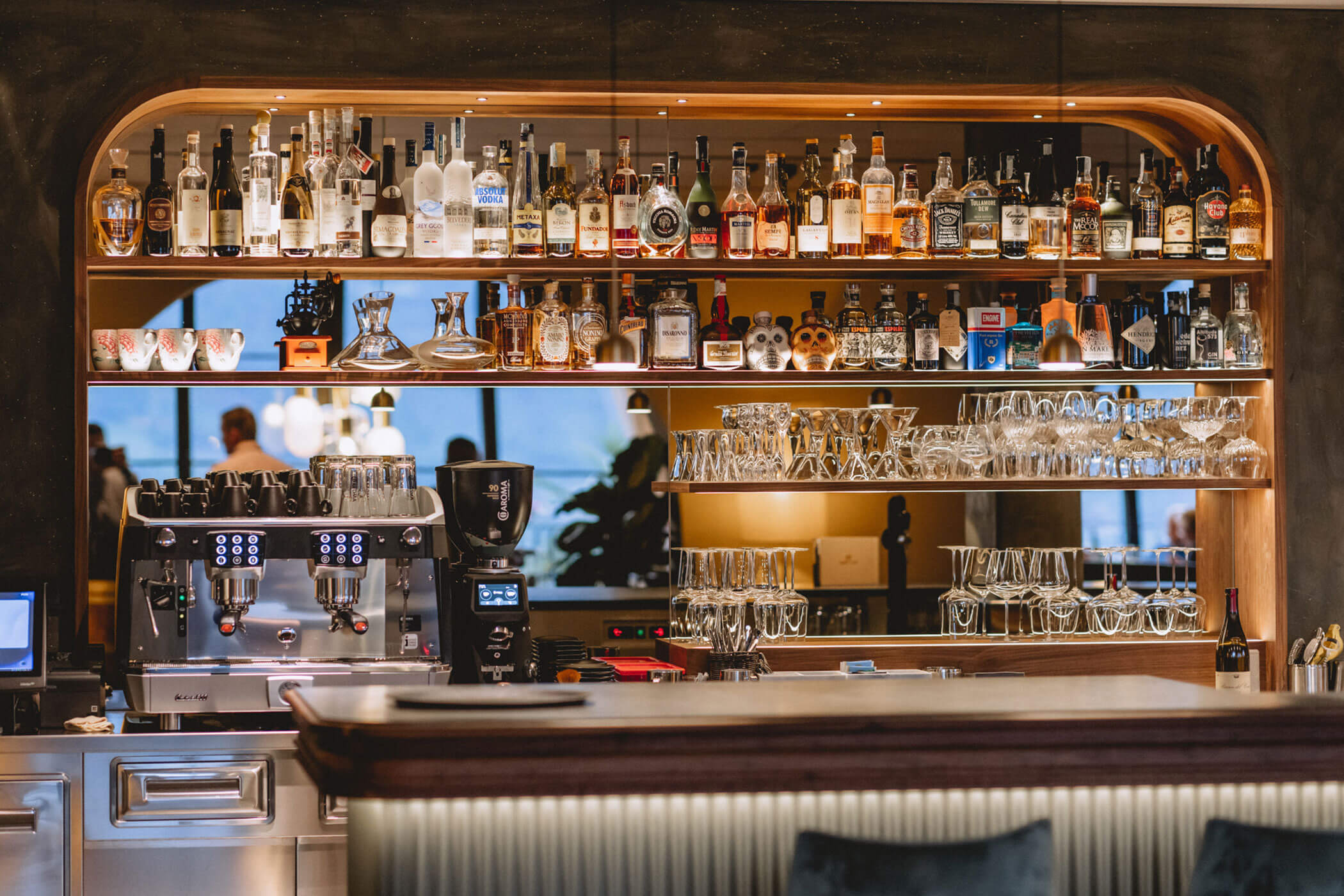 A stylish bar with shelves of liquor bottles, glassware, and a coffee machine behind a polished counter. - Hotel Lagrein