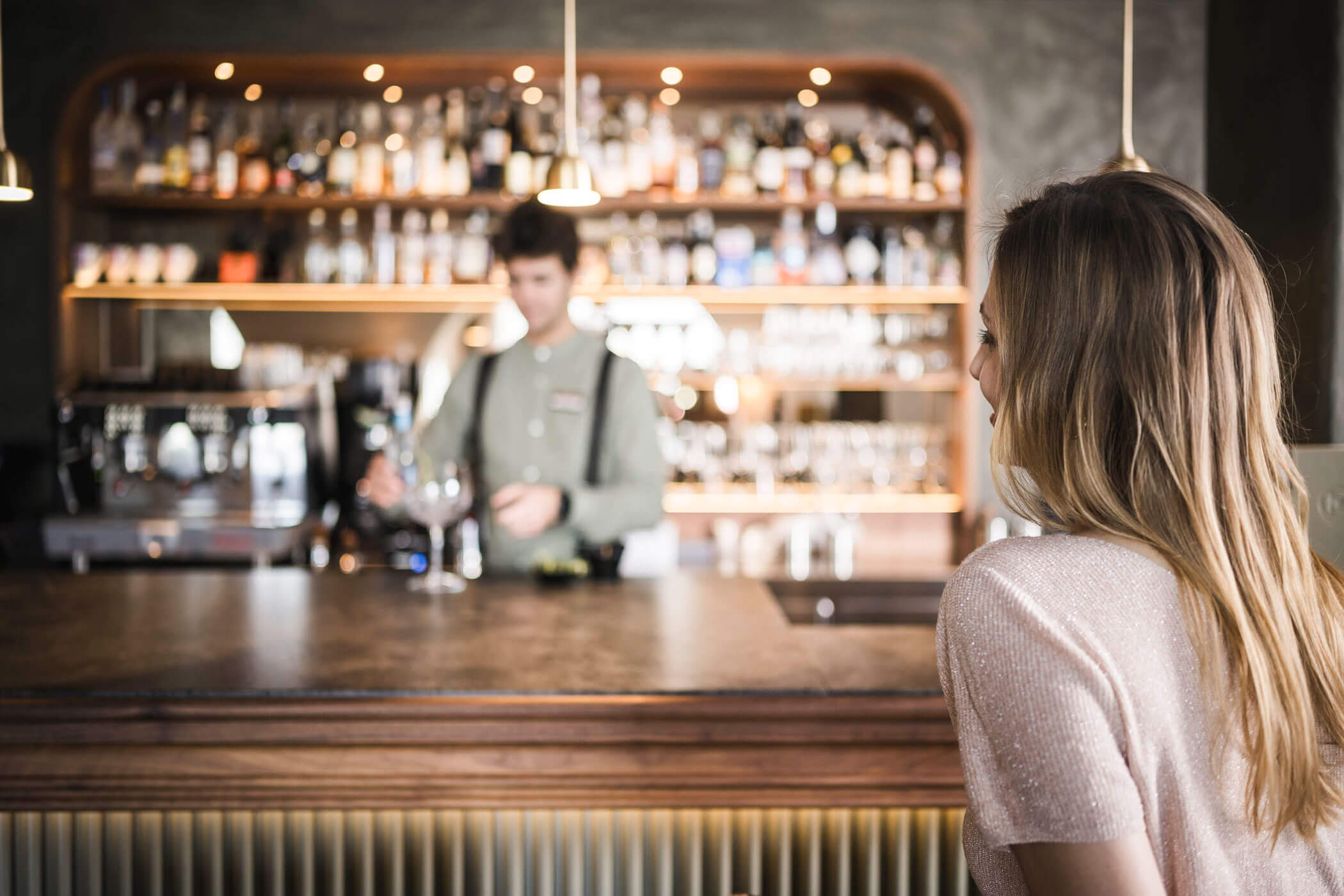 A woman sits at a bar counter while a bartender prepares drinks in the background. - Hotel Lagrein