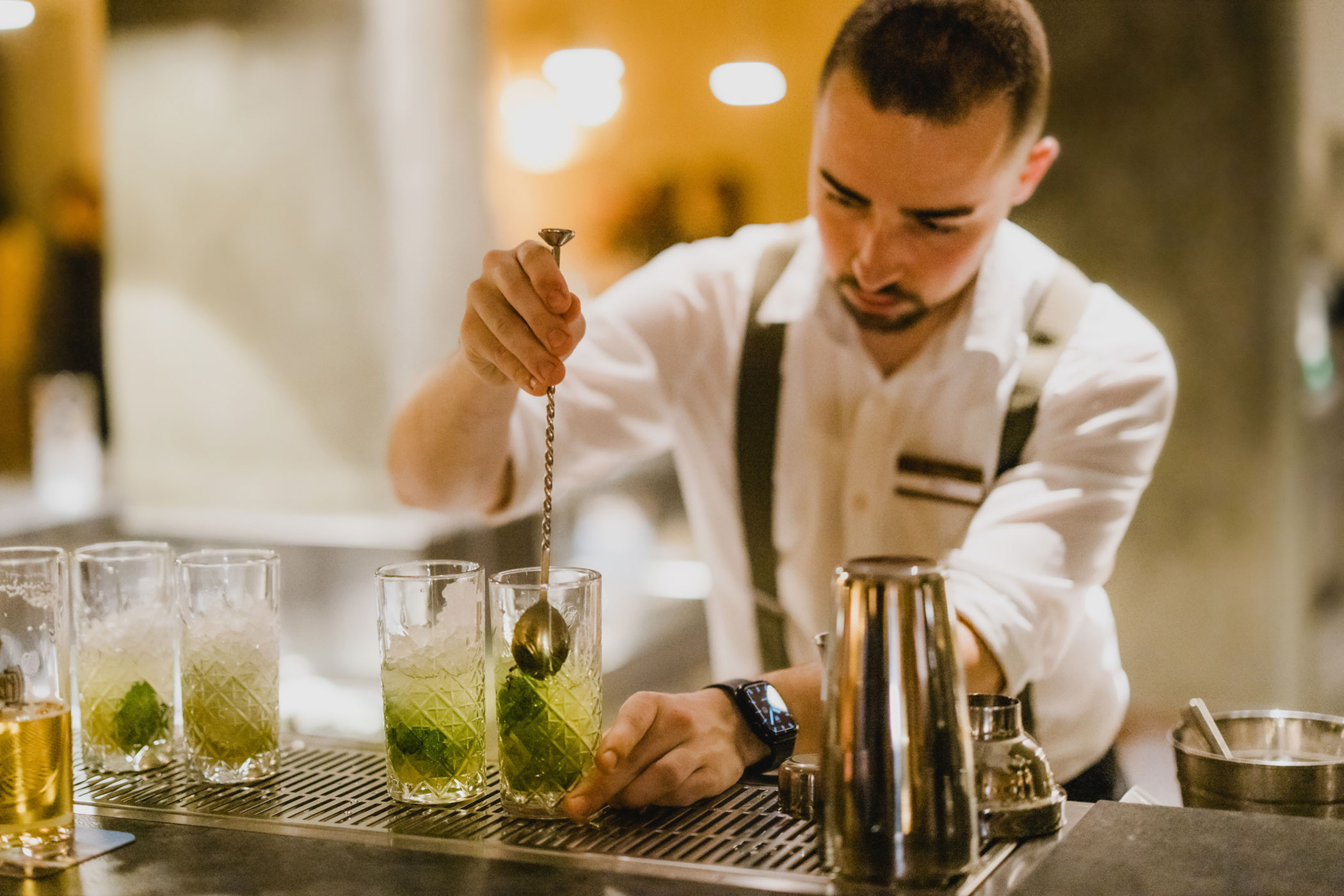 A bartender stirs a green cocktail in a glass at a bar, with other drinks and tools on the counter. - Hotel Lagrein