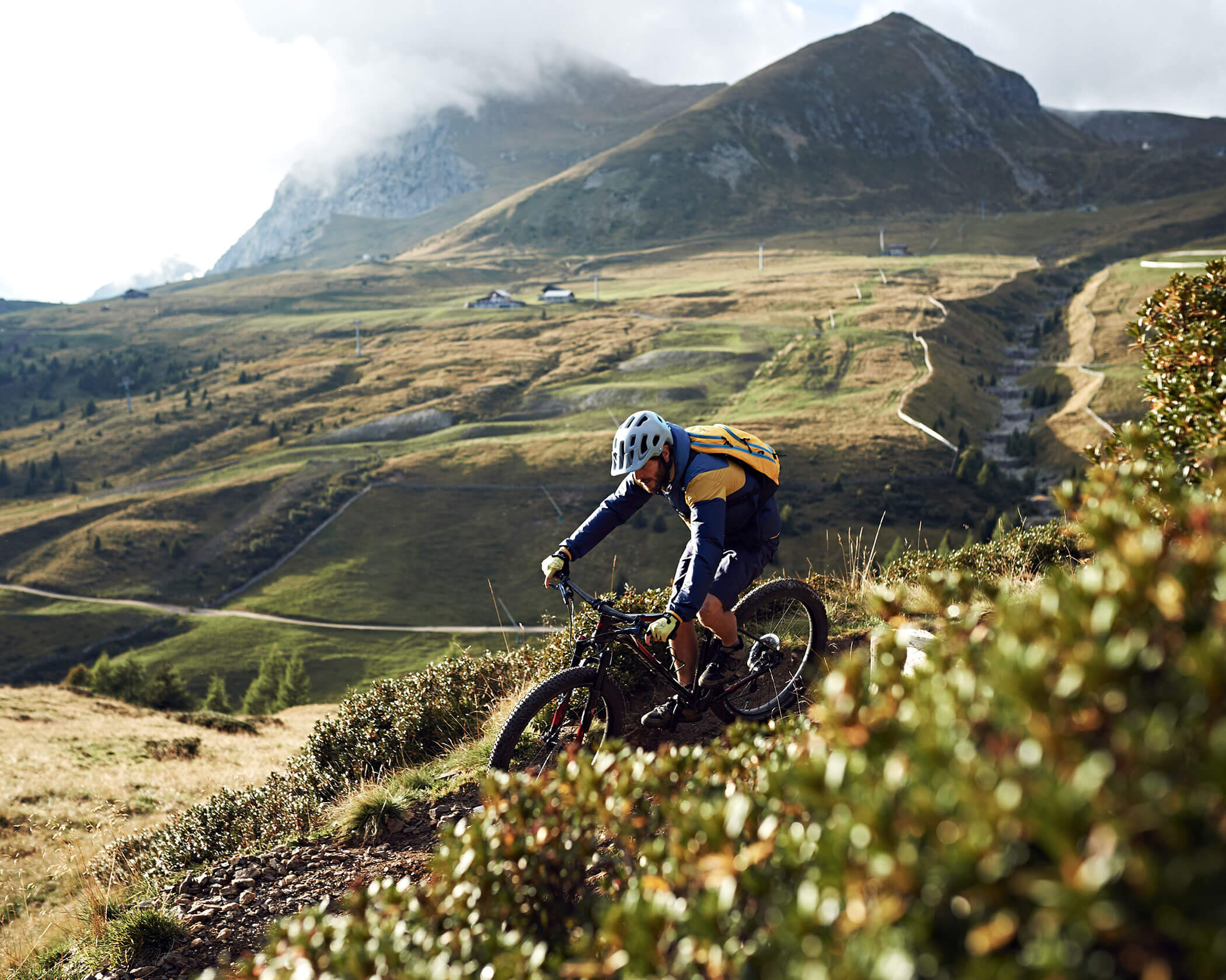 A cyclist rides a mountain bike down a rugged trail with hills and mountains in the background. - Hotel Lagrein