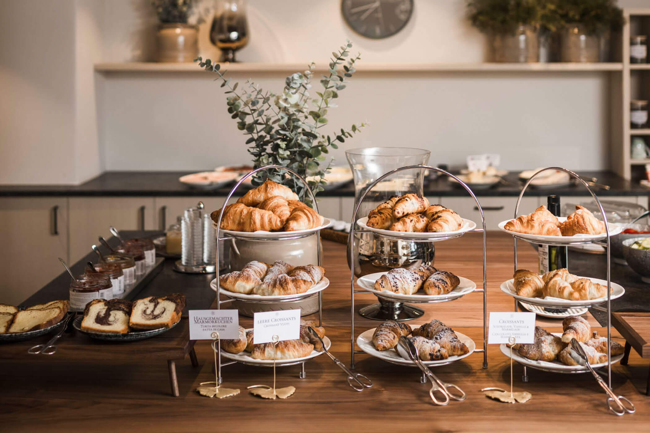 Three-tiered trays of assorted pastries and croissants on a wooden table in a modern kitchen setting. - Hotel Lagrein