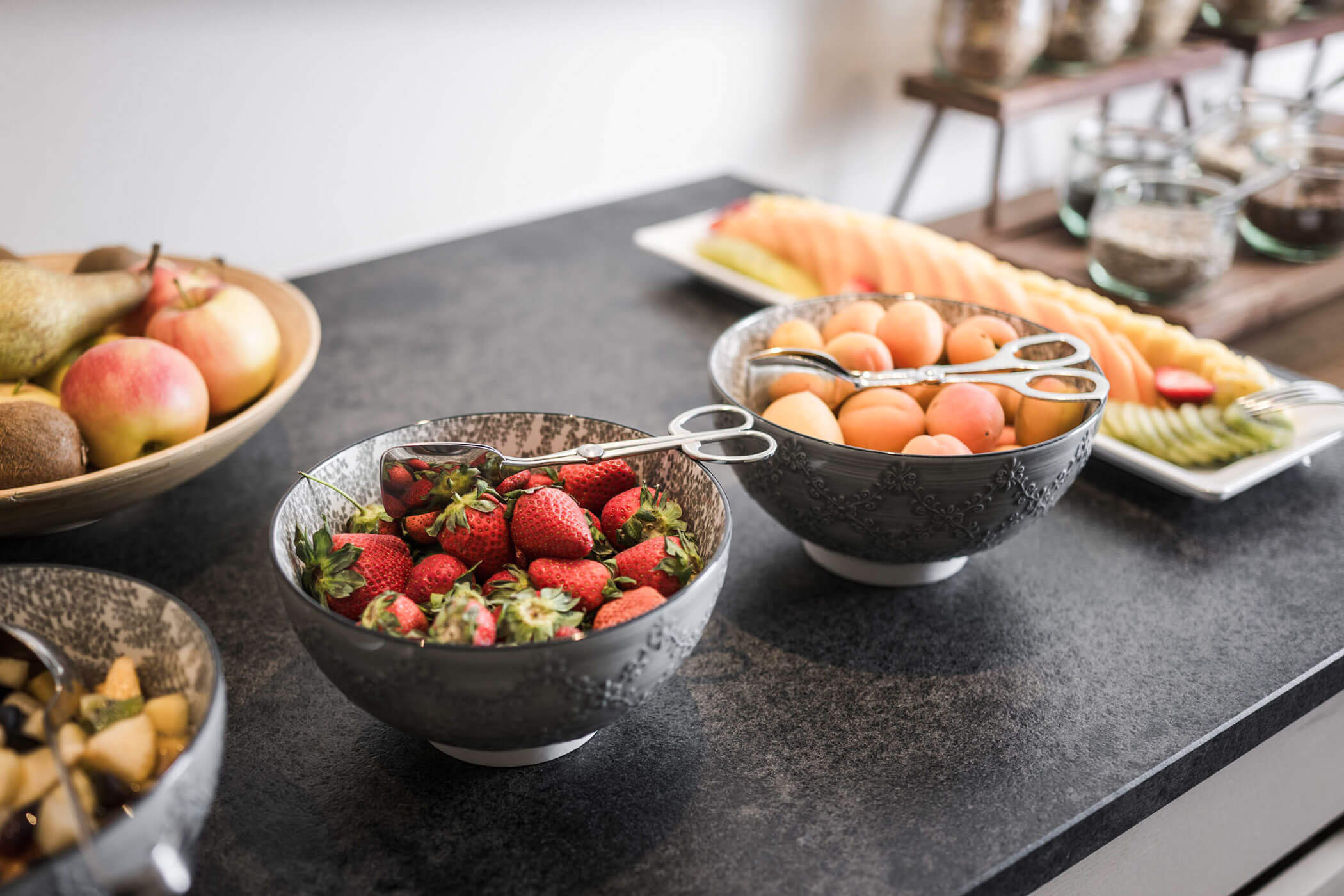 Bowls of fresh strawberries, apricots, and assorted fruit on a countertop, with serving utensils. - Hotel Lagrein