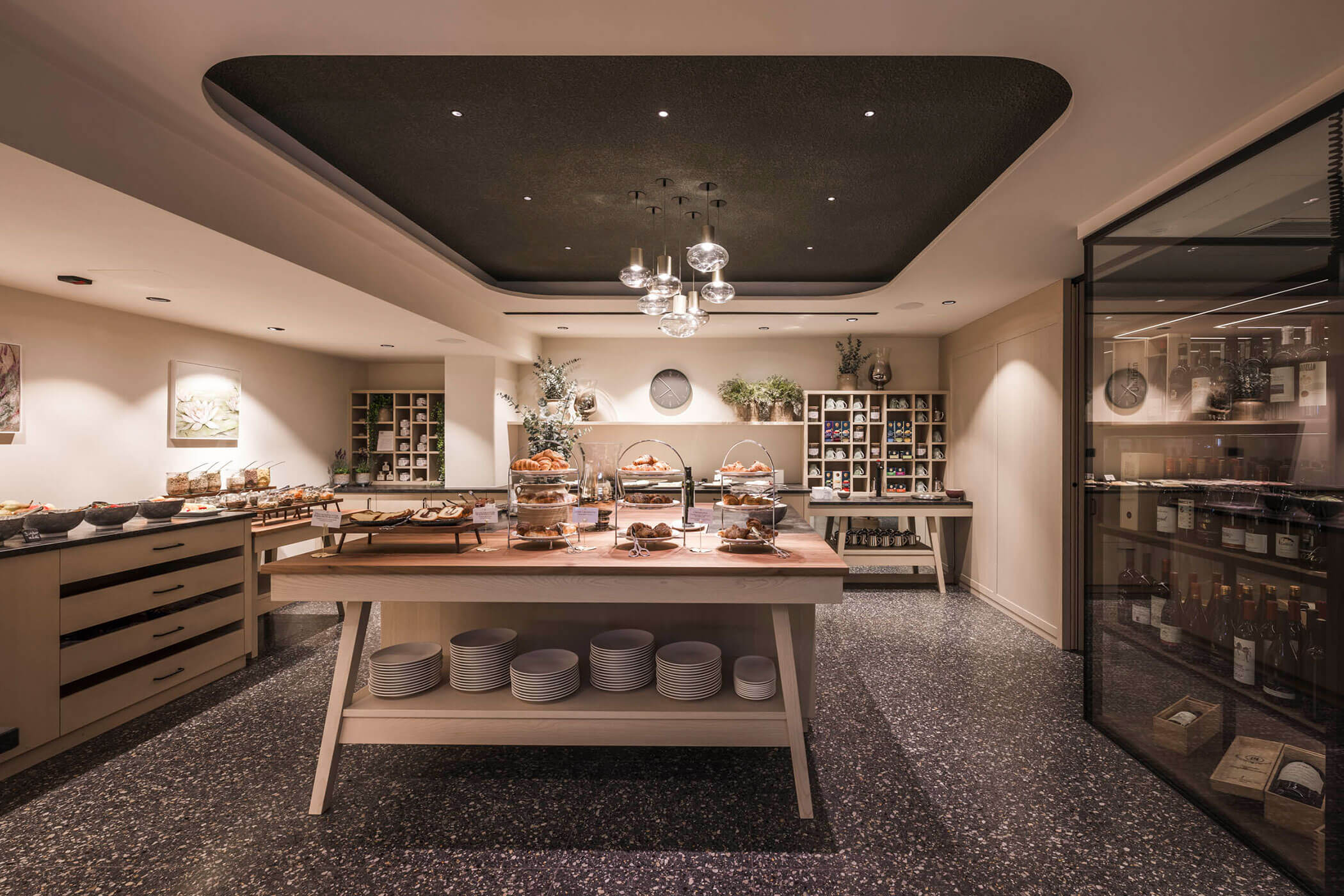 Modern buffet setup with pastries on a wooden table, shelves with jars, and a wine display on the right. - Hotel Lagrein