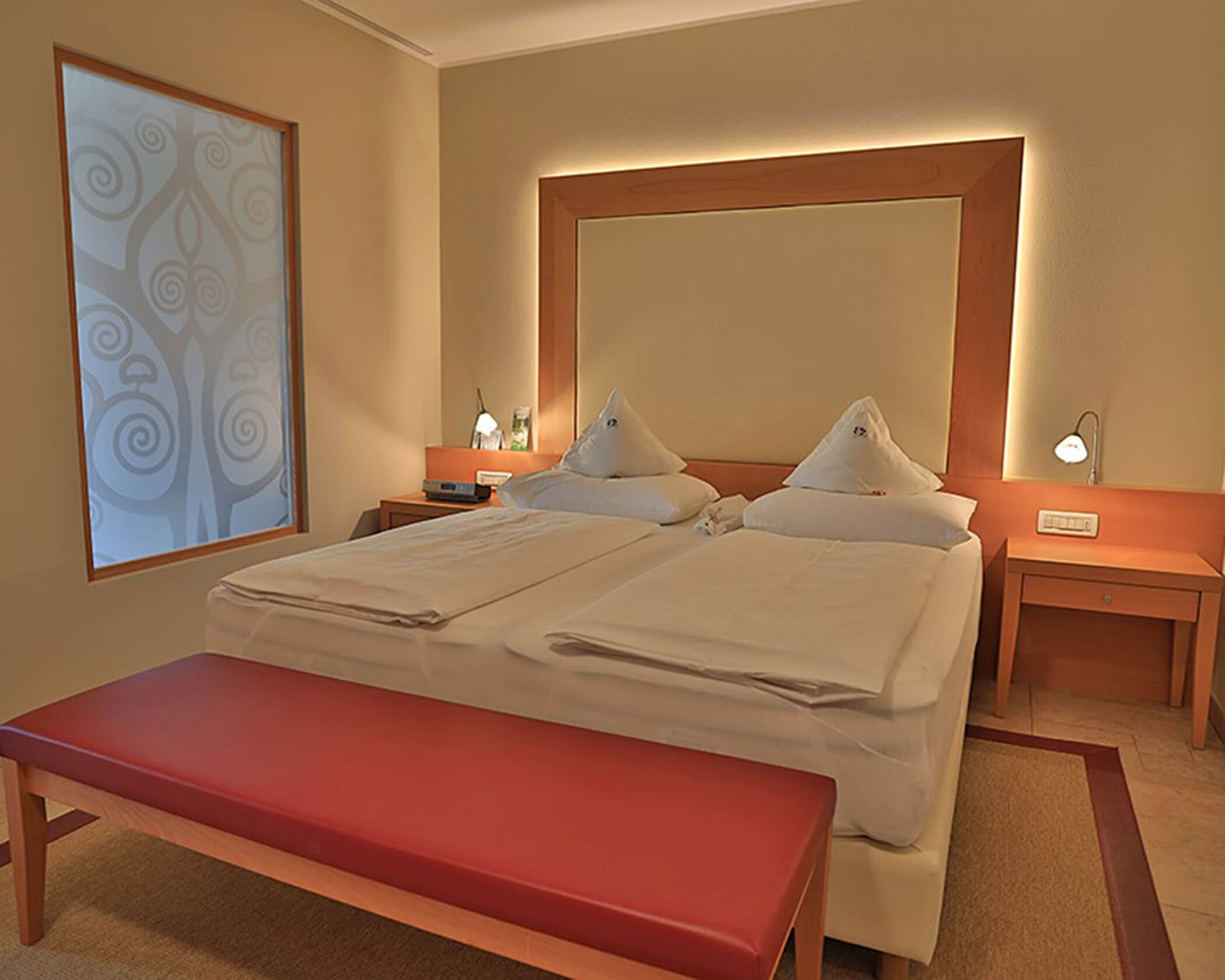 A hotel room with a double bed, red bench, bedside tables, and a decorative frosted glass window. - Hotel Lagrein