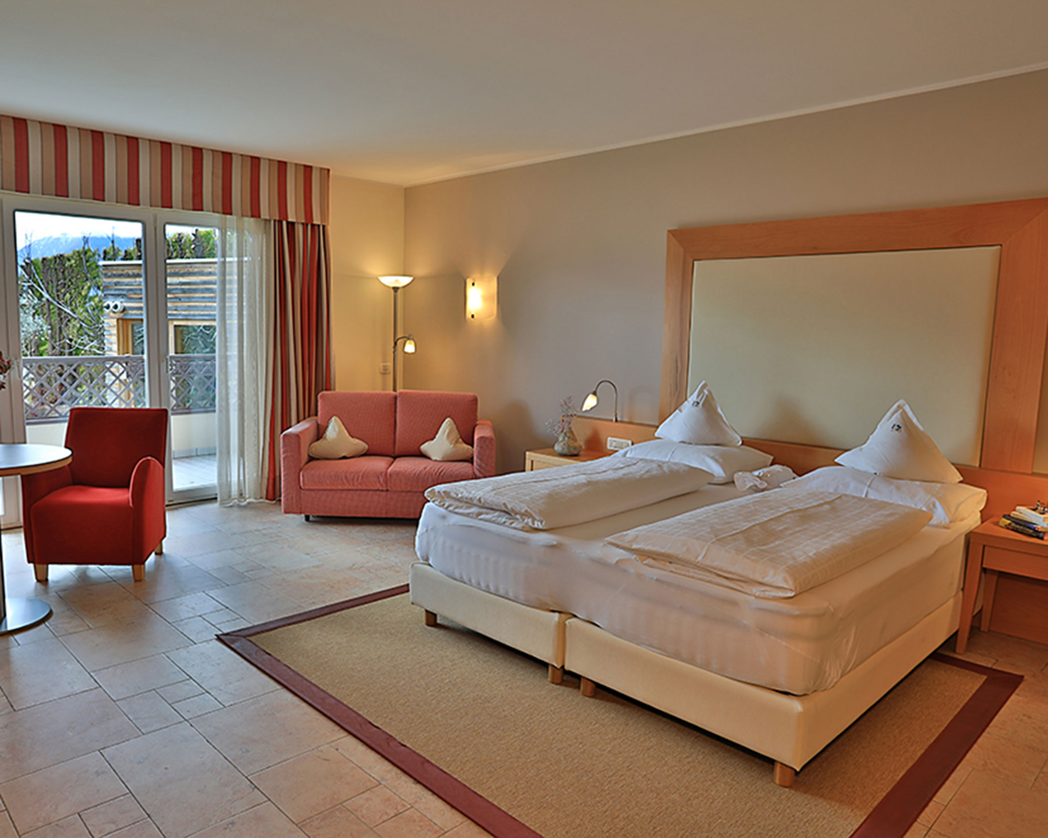 Spacious hotel room with twin beds, a red sofa, armchair, and a balcony with outdoor view. - Hotel Lagrein