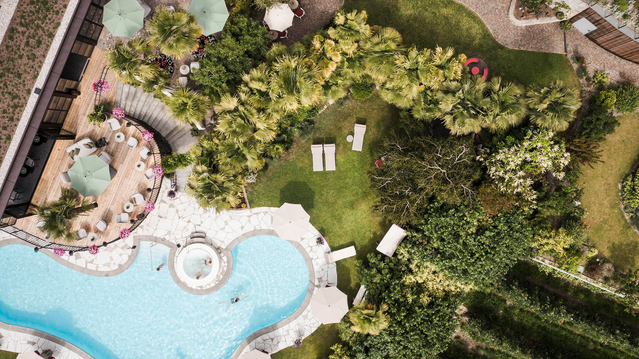 Aerial view of a pool area with lounge chairs, umbrellas, greenery, and people swimming. - Hotel Lagrein