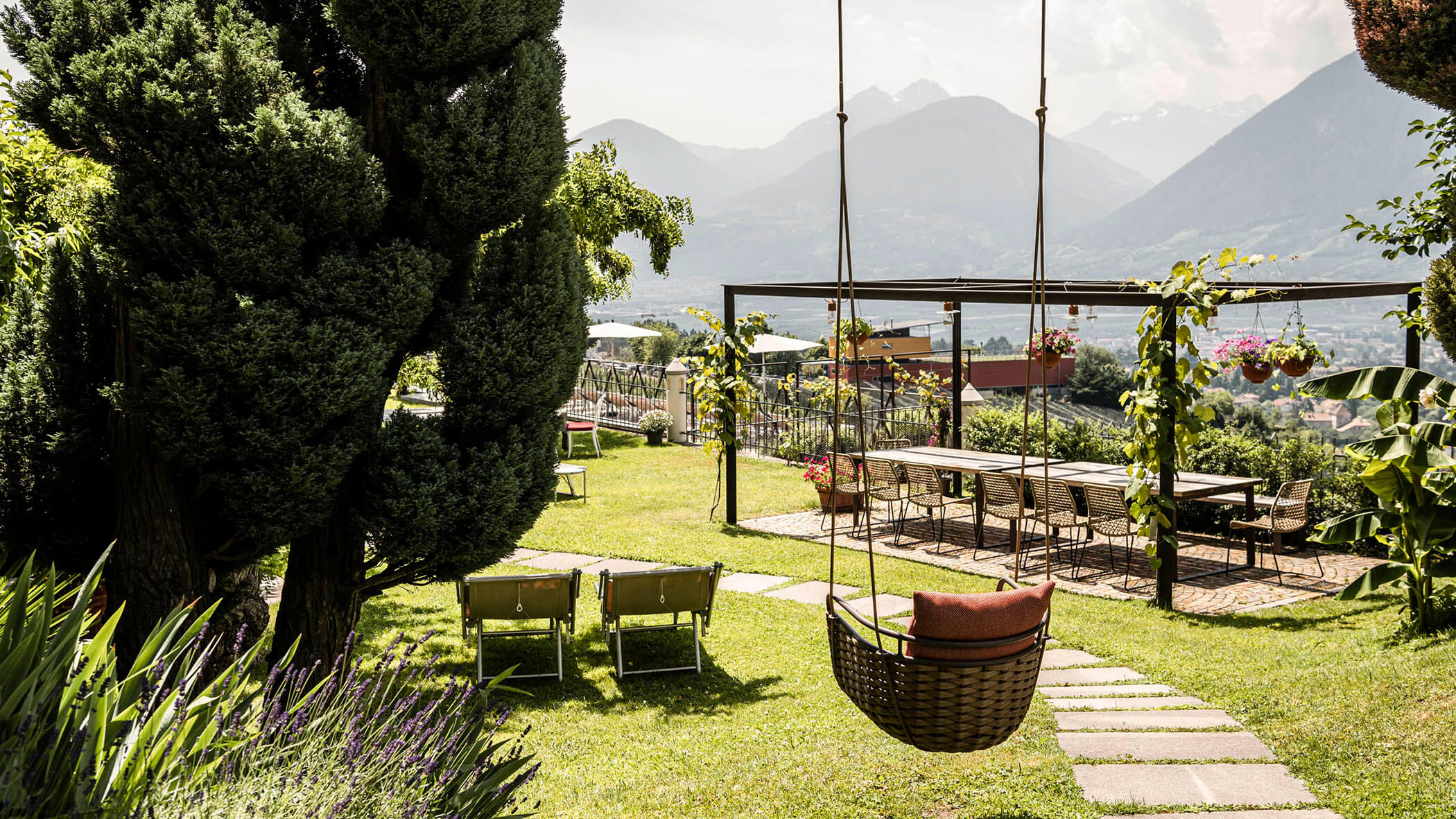 A garden with a hanging chair, lounge chairs, dining table, and mountains in the background. - Hotel Lagrein