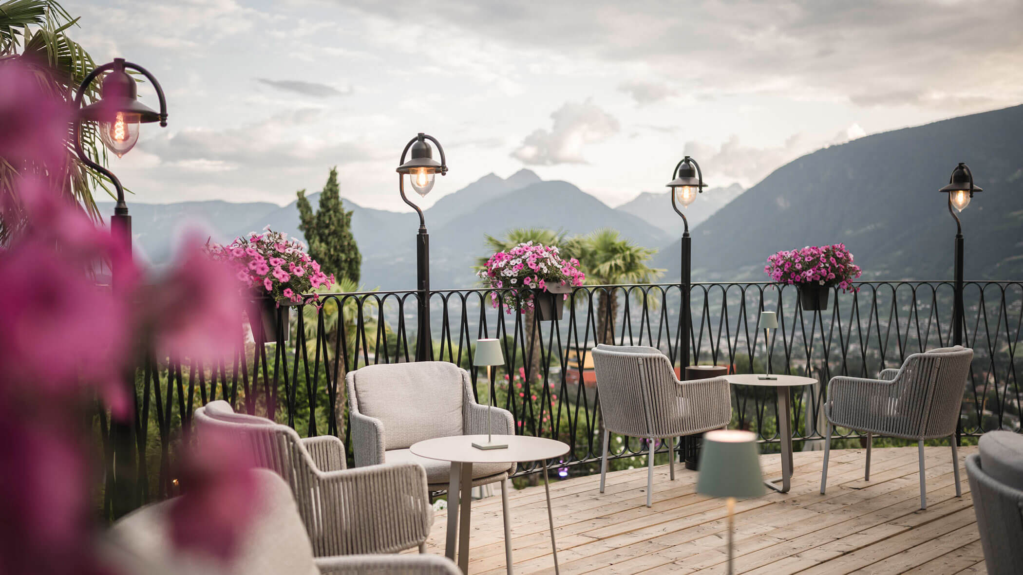 Outdoor terrace with cozy chairs, tables, and flowers, overlooking mountains under a cloudy sky. - Hotel Lagrein