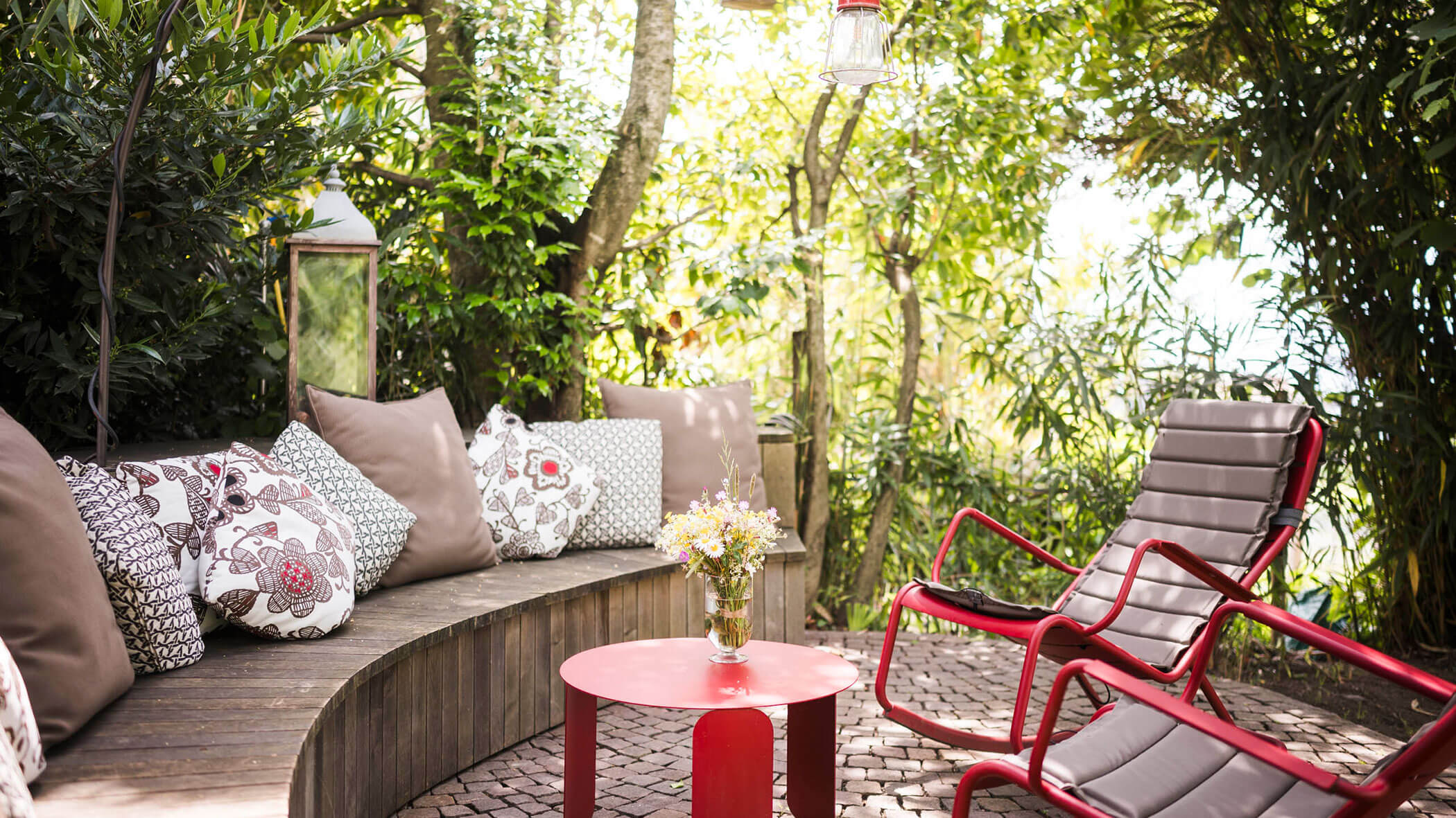 Cozy garden patio with curved bench, patterned cushions, red chairs, and a small red table with flowers. - Hotel Lagrein