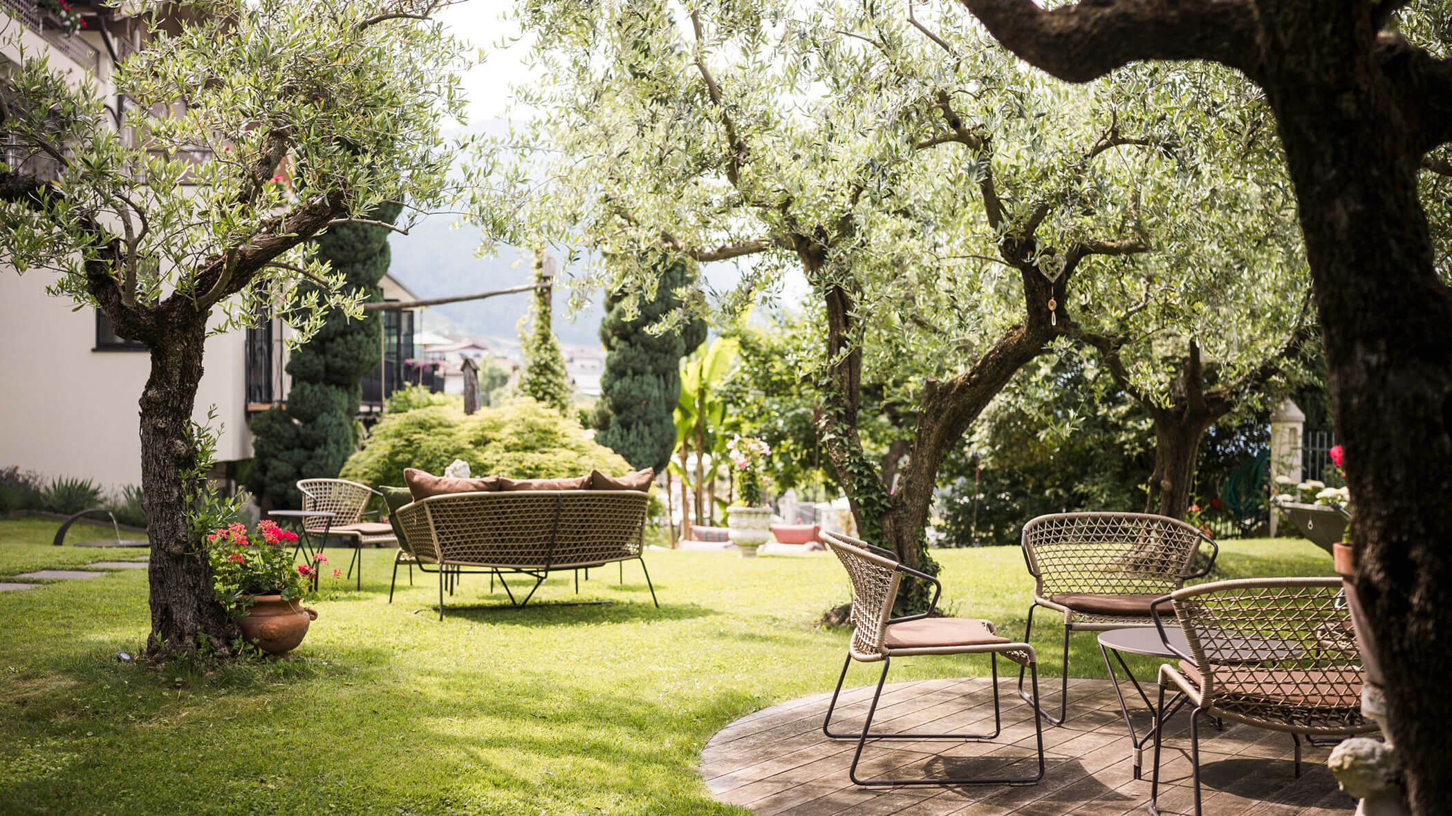 Wicker chairs and tables arranged under leafy trees in a sunny, green garden. - Hotel Lagrein