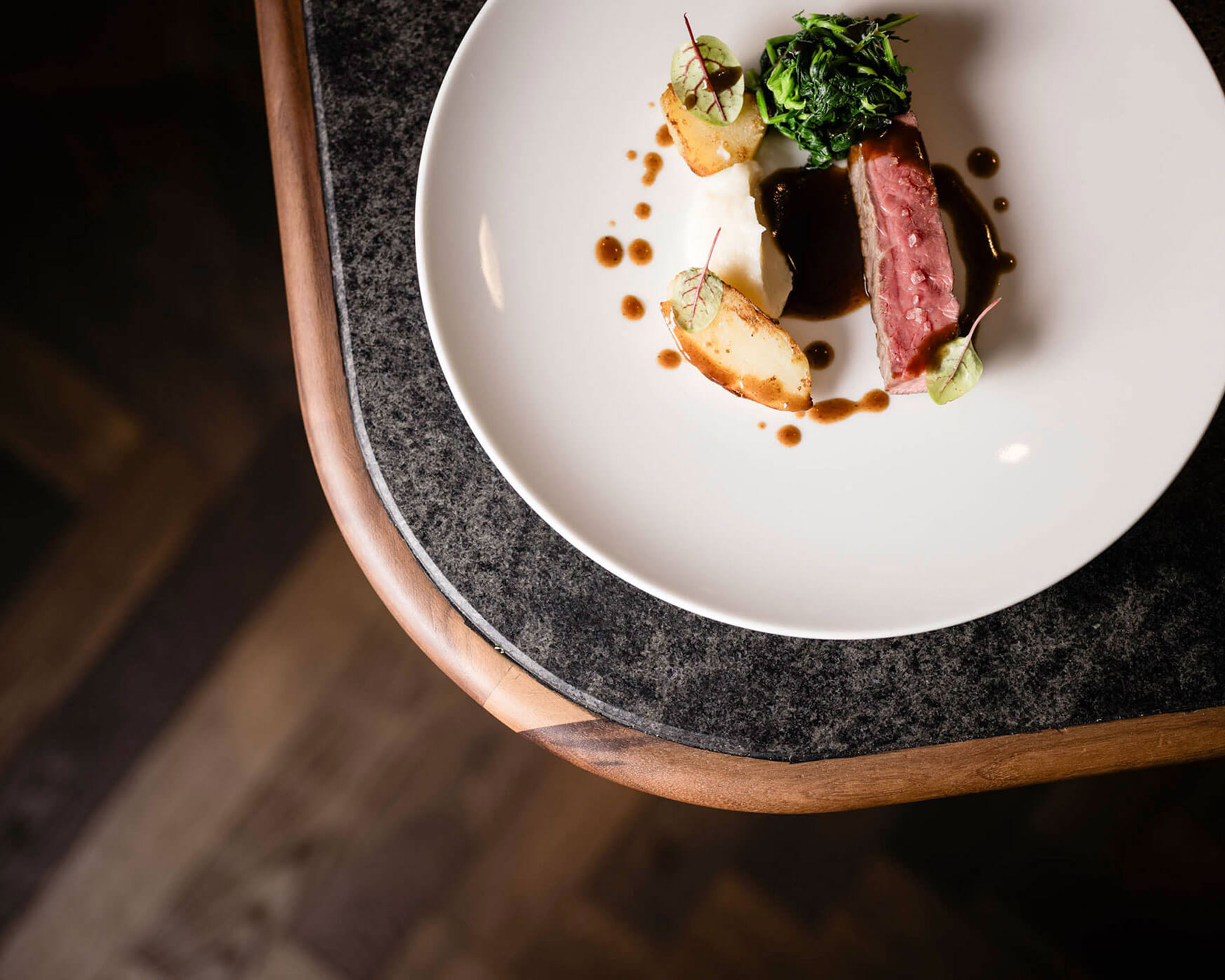 A gourmet dish with sliced meat, potatoes, greens, and sauce artfully plated on a white round plate. - Hotel Lagrein