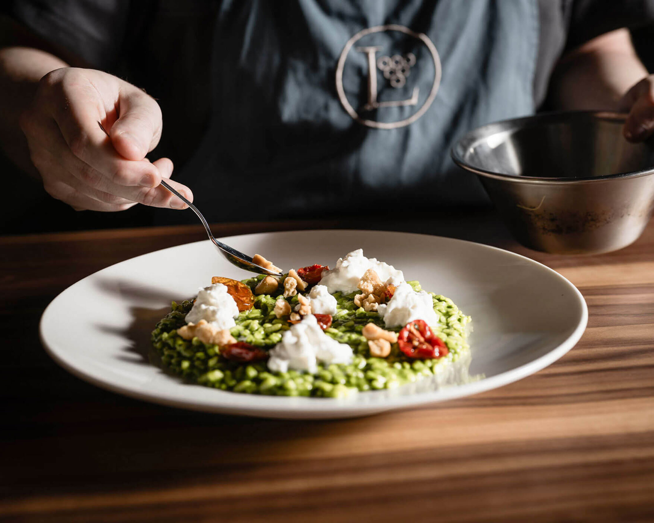 A person adds toppings to a plate of green risotto with tomatoes, nuts, and cheese on a wooden table. - Hotel Lagrein