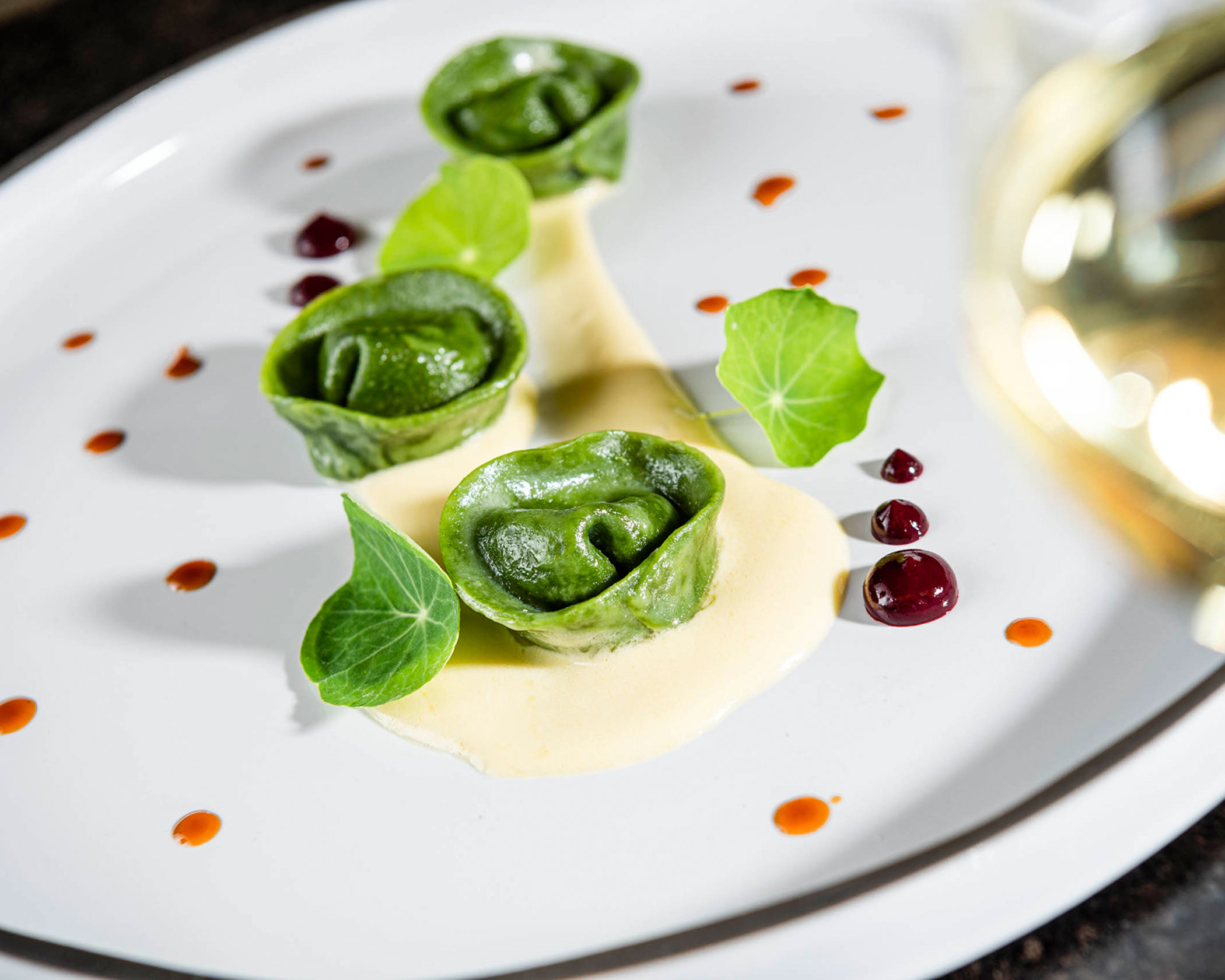 Three green tortellini with sauce, garnished with edible leaves and dots of red and orange sauce on a white plate. - Hotel Lagrein