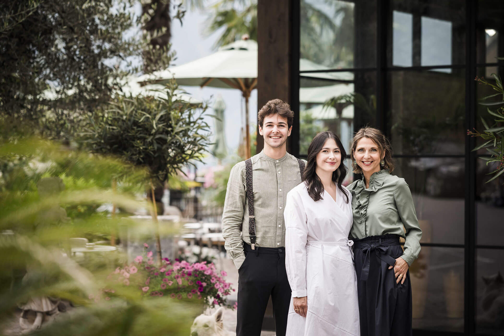 Three people stand smiling together outdoors near a building with greenery and patio umbrellas in the background. - Hotel Lagrein