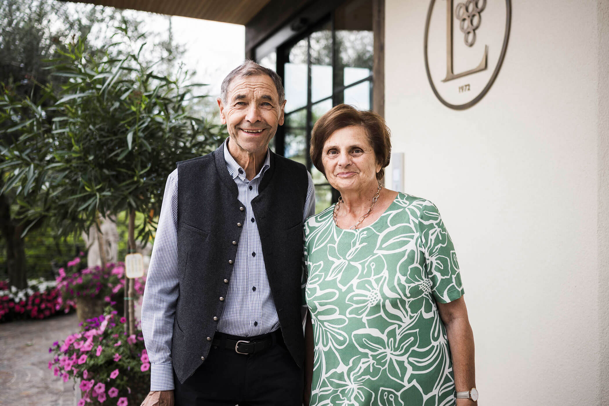 Older man and woman smiling, standing together outside a building with flowers and plants nearby. - Hotel Lagrein