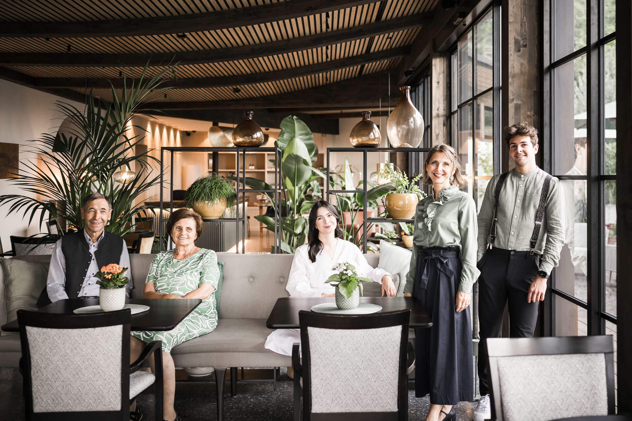 Five people in a modern, bright restaurant with plants and large windows, smiling at the camera. - Hotel Lagrein