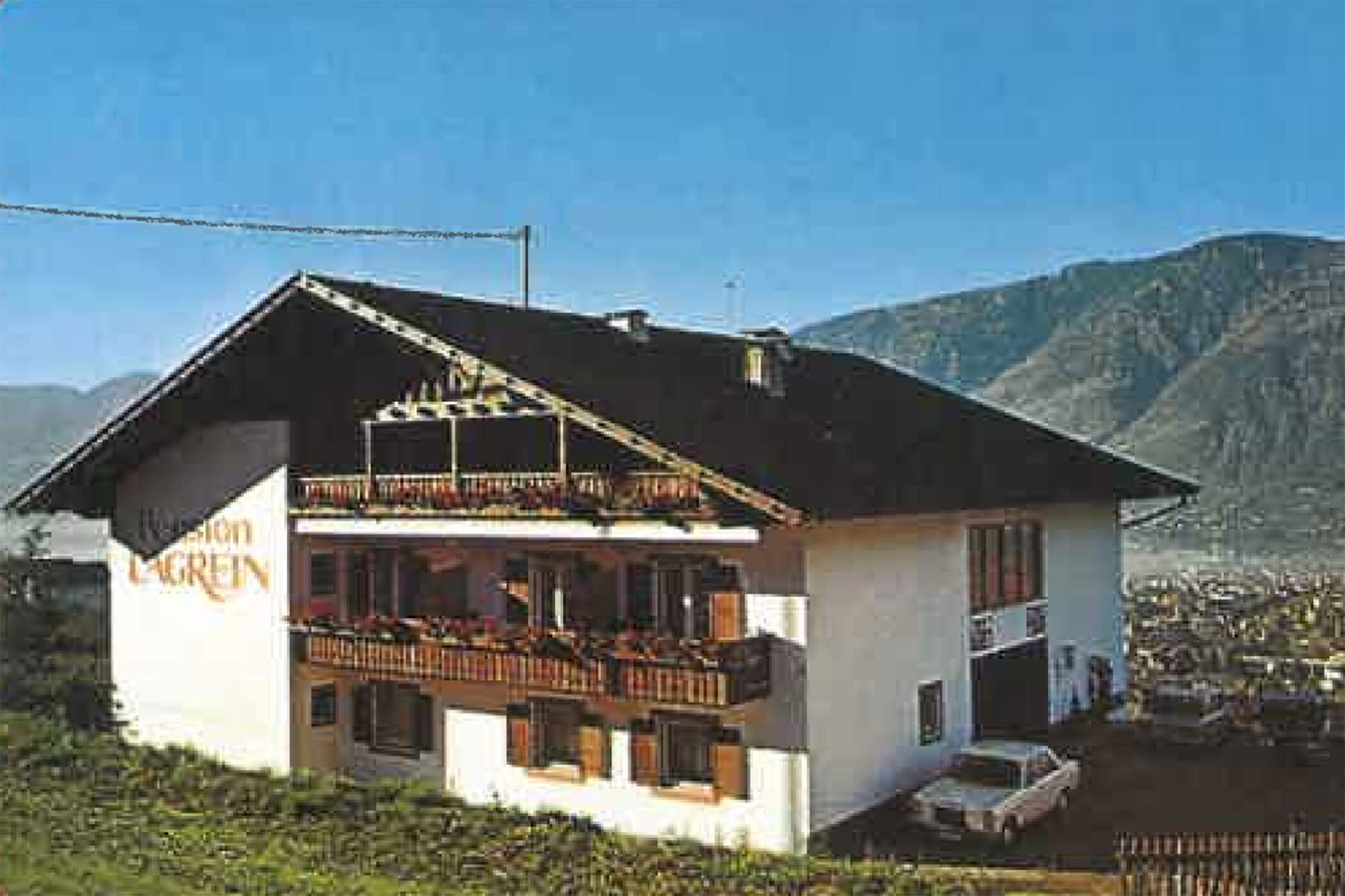 A white house with balconies, mountains in the background, and a car parked outside on a clear day. - Hotel Lagrein