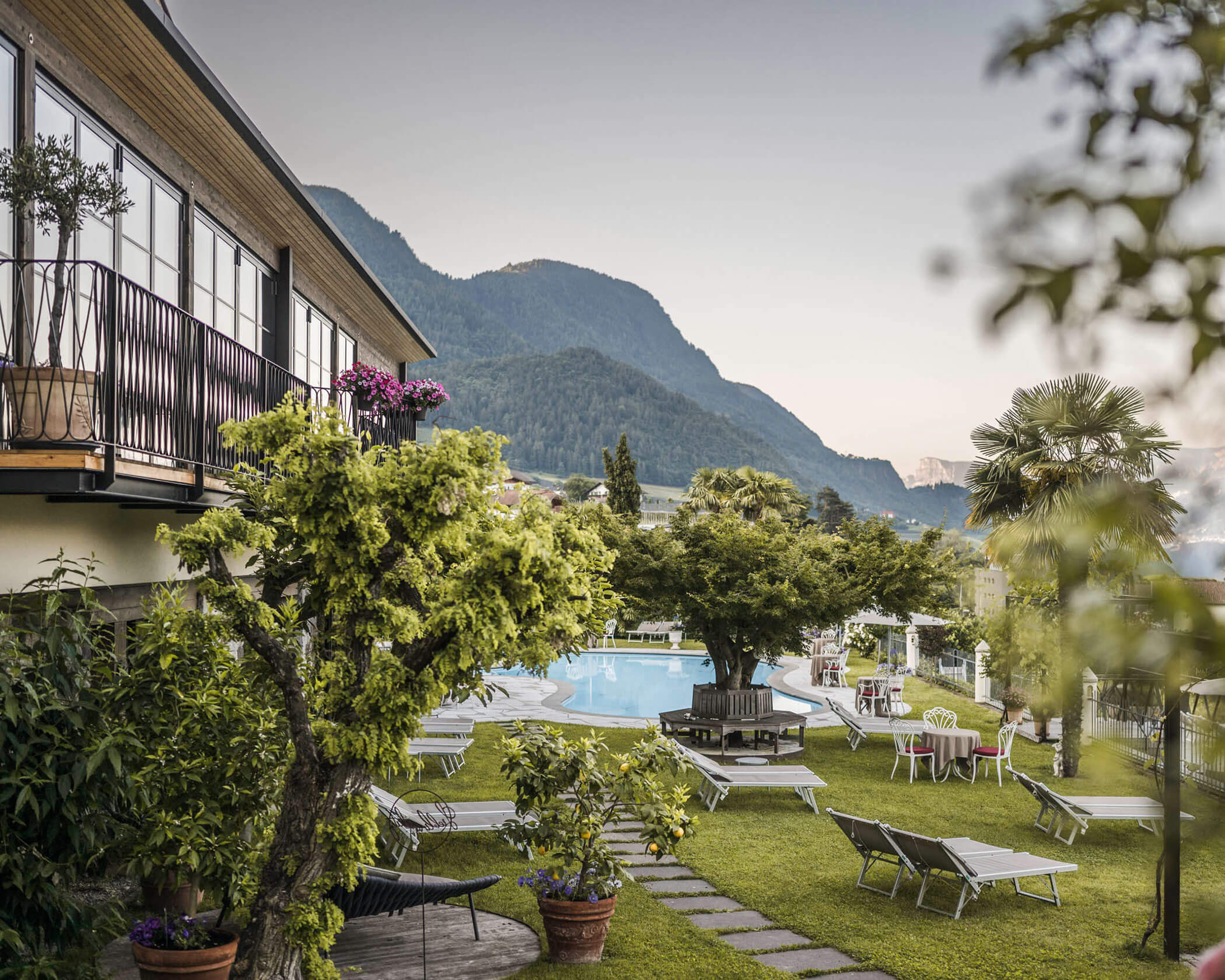 Lush garden with lounge chairs and pool, surrounded by mountains and a modern building at sunset. - Hotel Lagrein