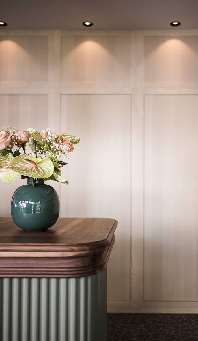 A green vase with flowers sits on a wooden counter against a light-paneled wall with spotlights above. - Hotel Lagrein