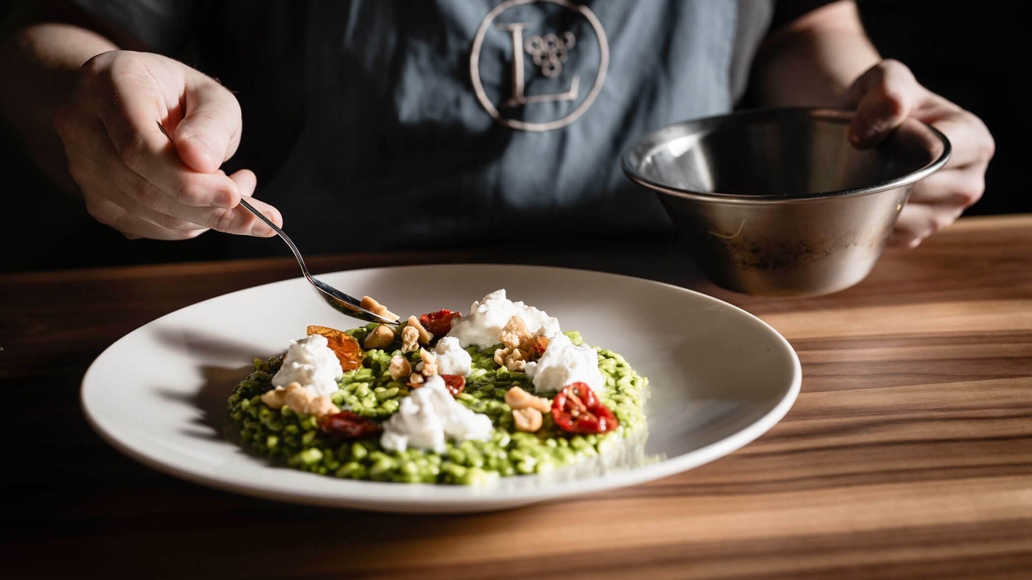 Person plating a green risotto dish with cheese, walnuts, and roasted tomatoes on a wooden table. - Hotel Lagrein