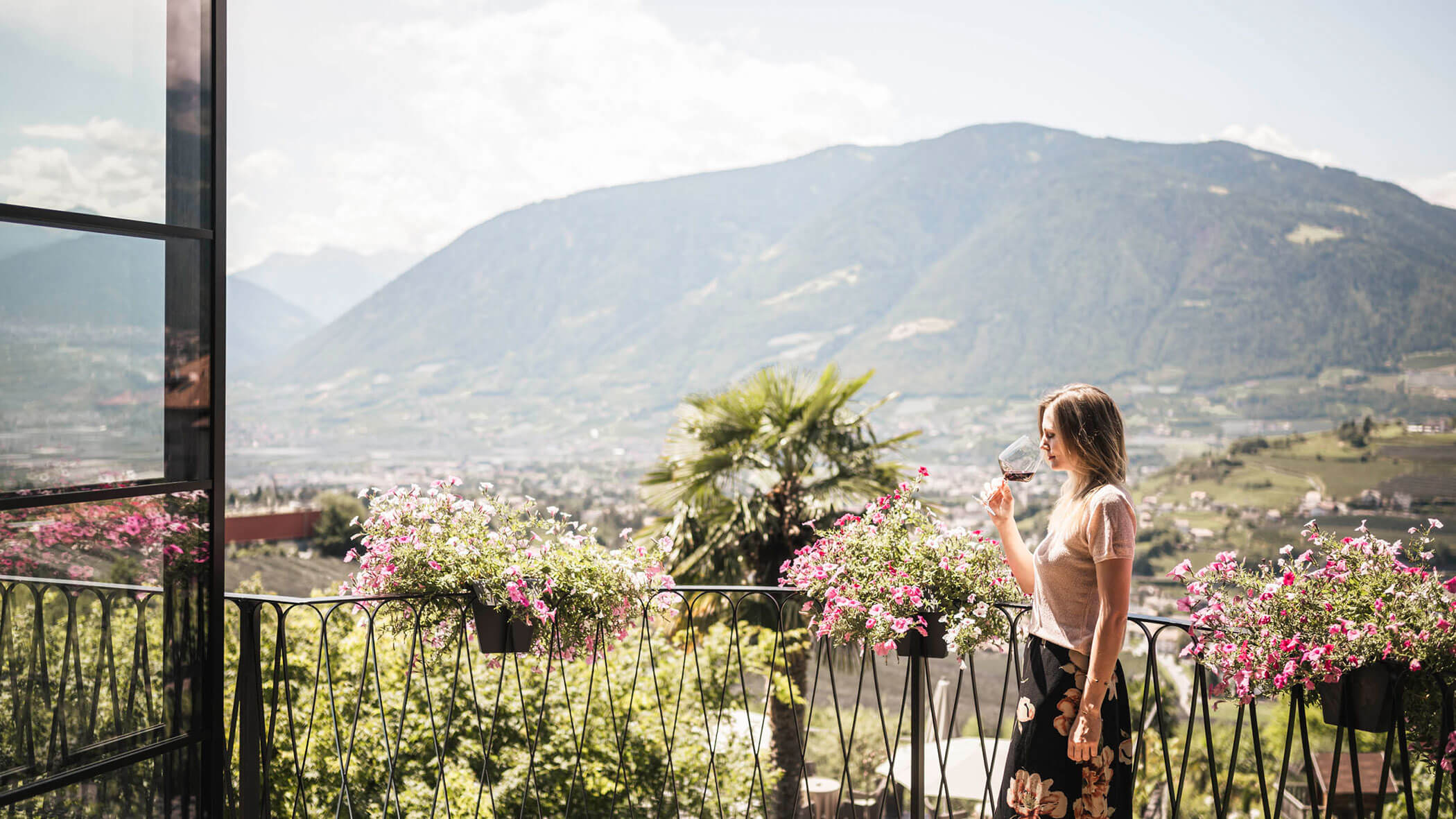 Woman drinks wine on a balcony with pink flowers, overlooking a scenic mountain and valley landscape. - Hotel Lagrein