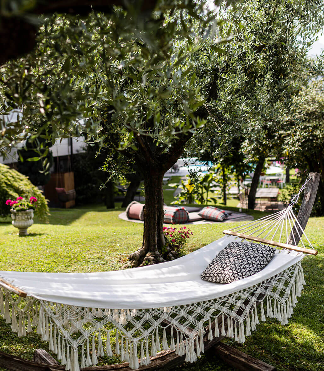A white hammock with a patterned pillow hangs between trees in a lush, sunny garden. - Hotel Lagrein