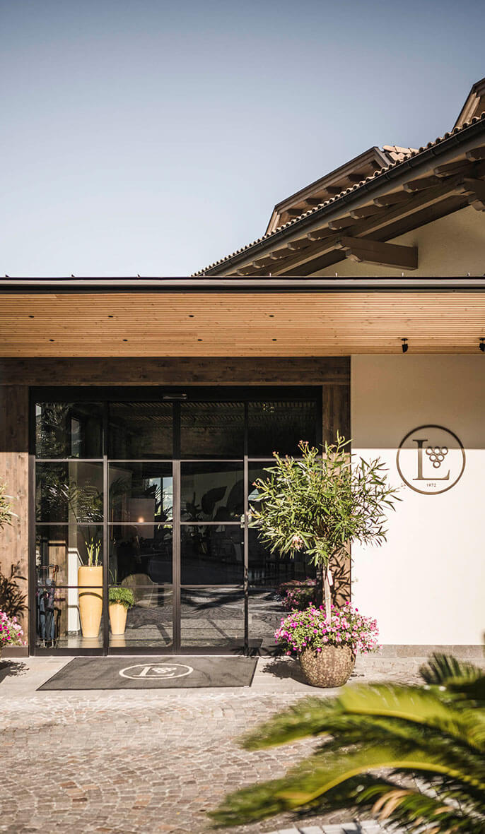 Modern building entrance with glass doors, potted plants, and a round wall logo featuring a grape cluster. - Hotel Lagrein