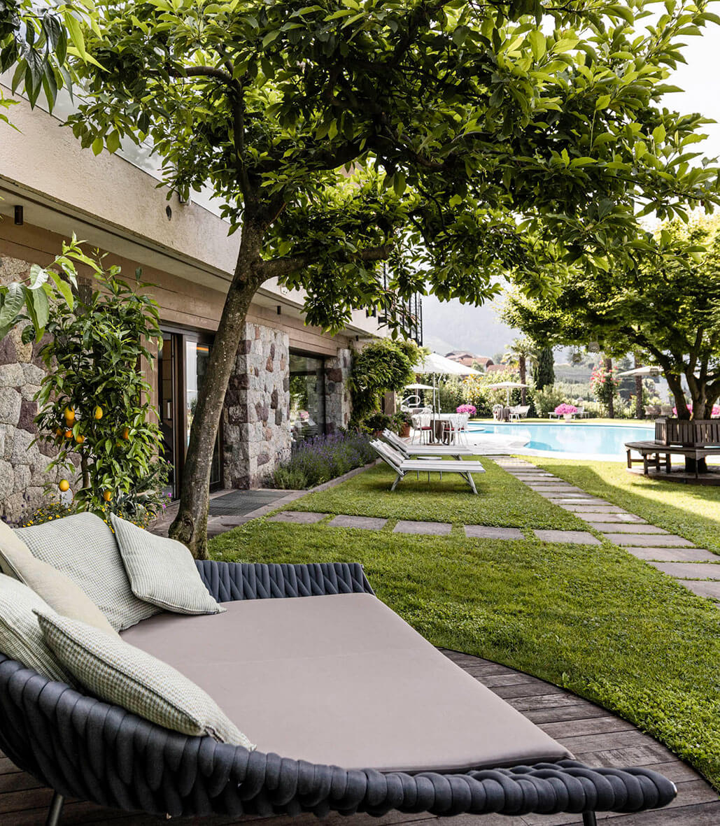 Outdoor lounge area with a cushioned daybed, trees, stone house, and pool in the background. - Hotel Lagrein